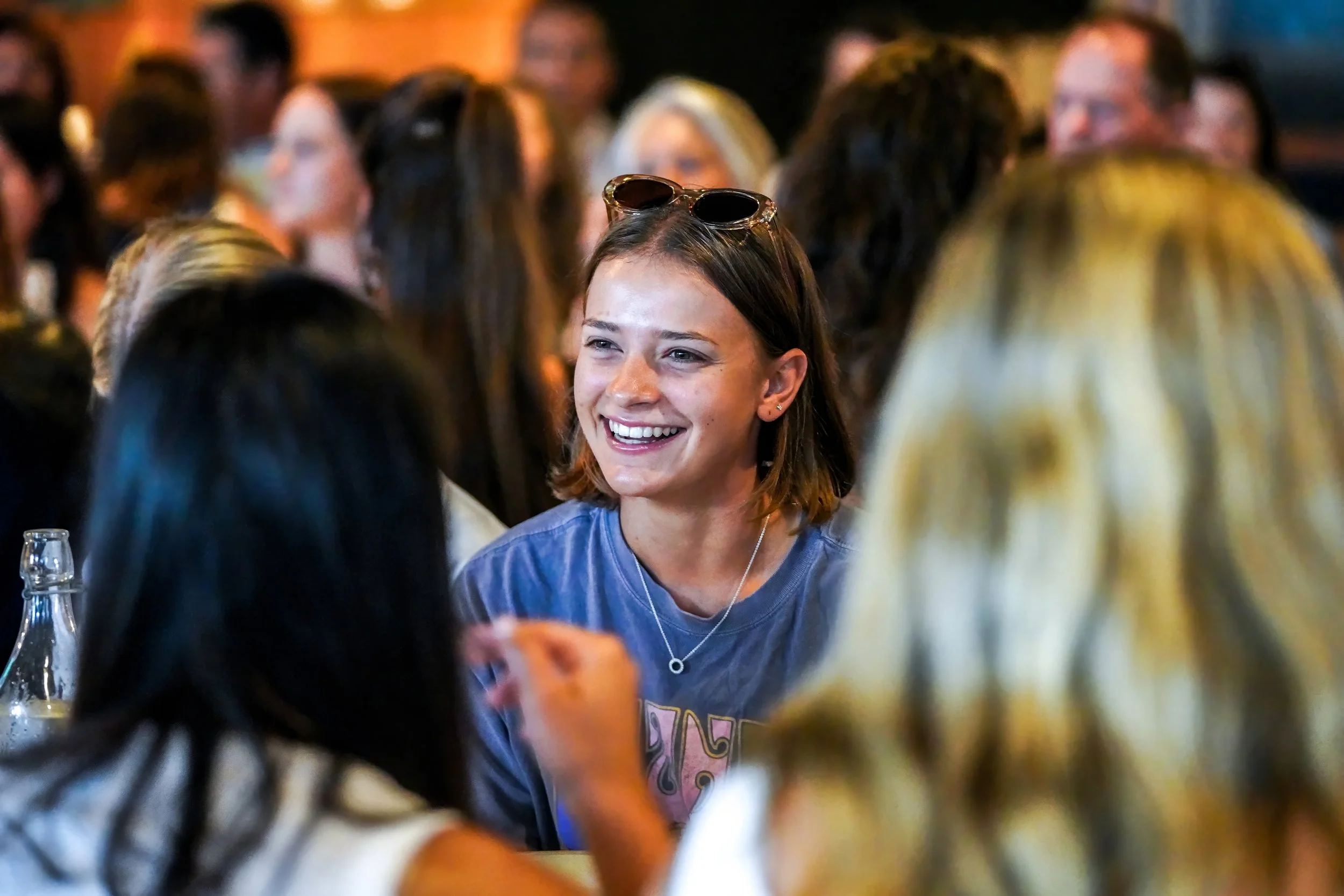A young woman with shoulder-length brown hair, sunglasses on her head, smiling and talking with friends at a crowded indoor event.