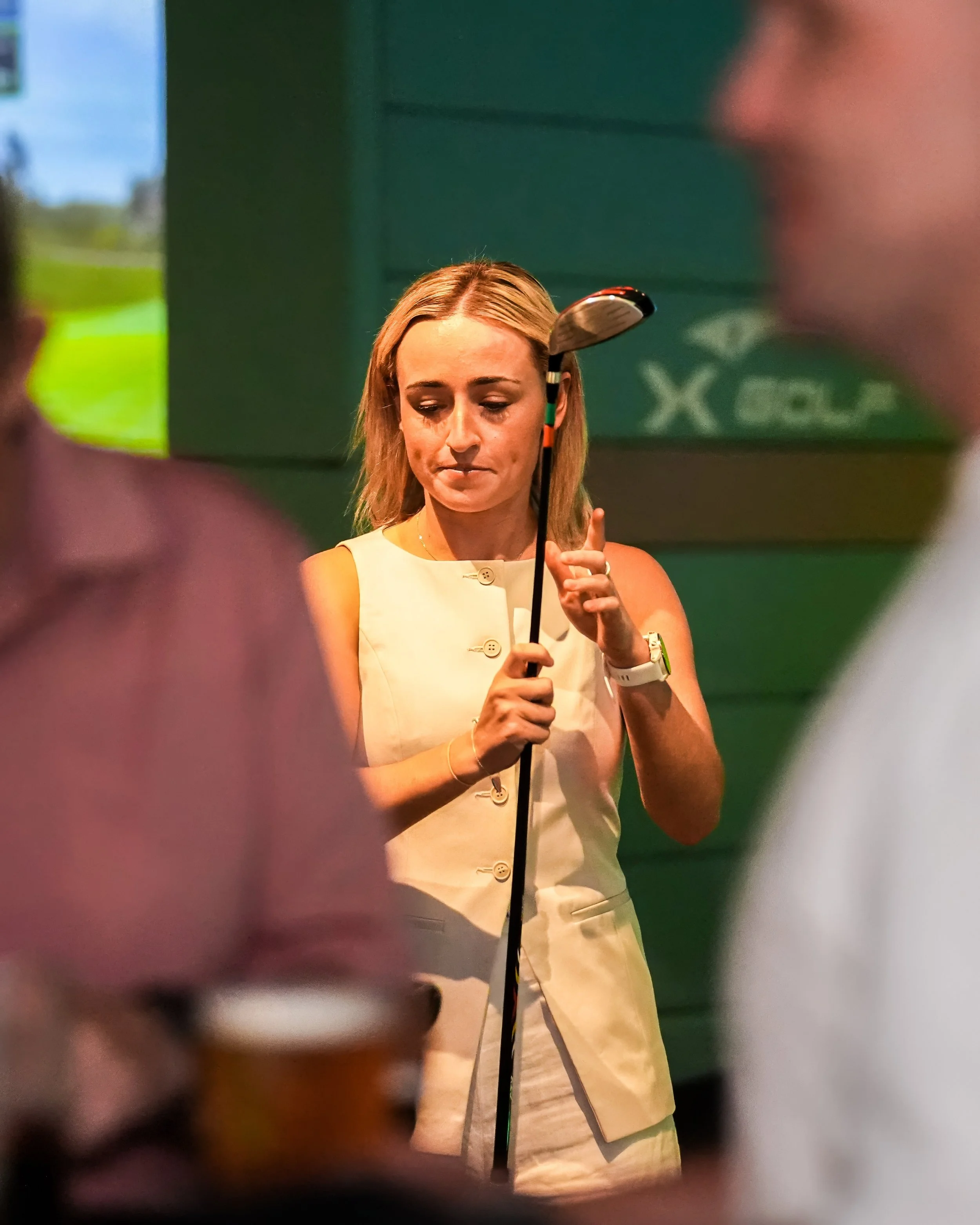 A woman in a cream-colored sleeveless dress holding a golf club, looking down thoughtfully, in a golf-themed setting with blurred figures nearby.