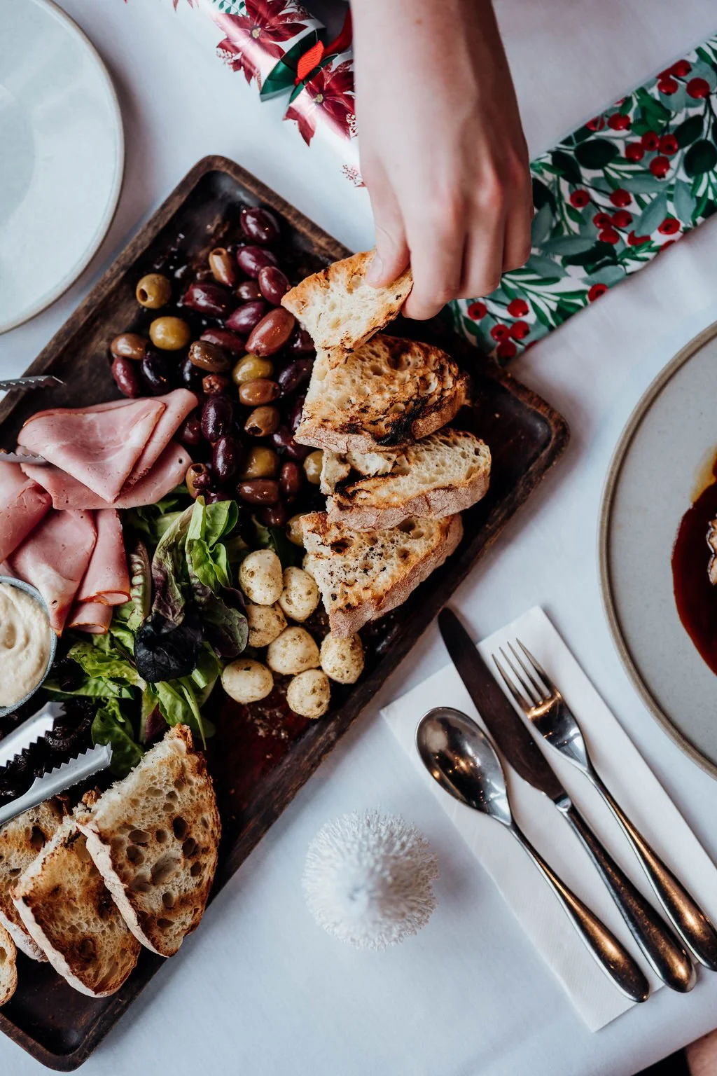 A person holding a piece of bread over a charcuterie board with grapes, olives, ham, salad greens, bread slices, and cheese. Tableware and utensils are arranged nearby.