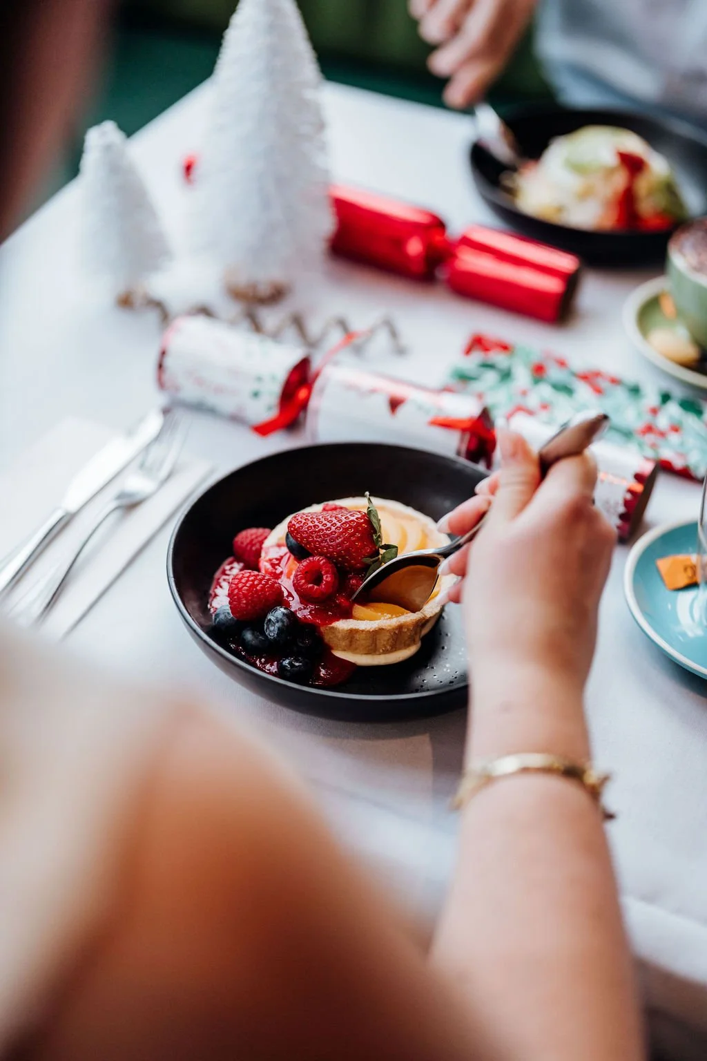 A person holding a spoon over a bowl of toast topped with mixed berries and a fried egg on a festive table surrounded by Christmas decorations.