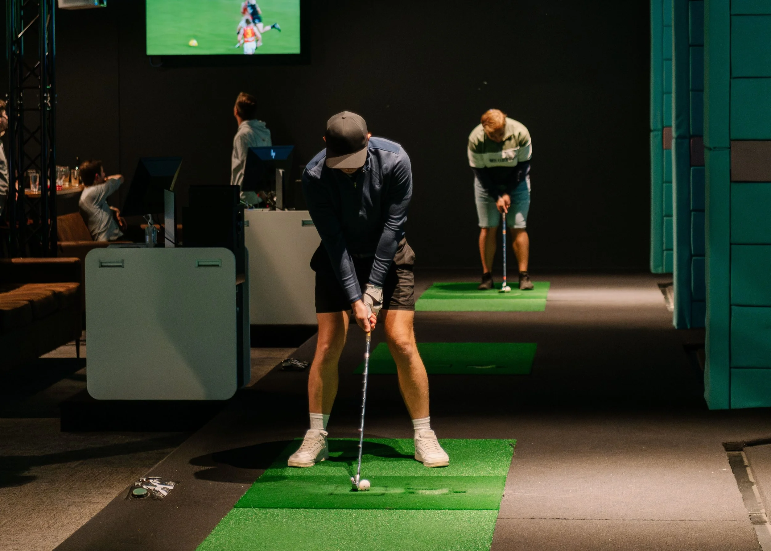 Person wearing a Santa hat practicing putting in an indoor golf simulator, with two women watching, also wearing Santa hats.