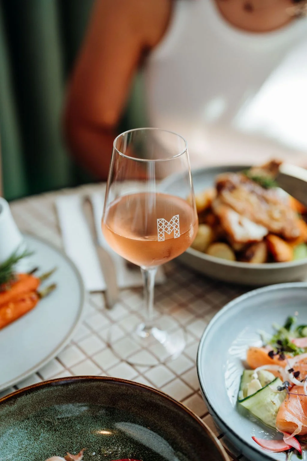 A glass of rosé wine with a geometric 'M' logo on it, surrounded by plates of various dishes on a table.