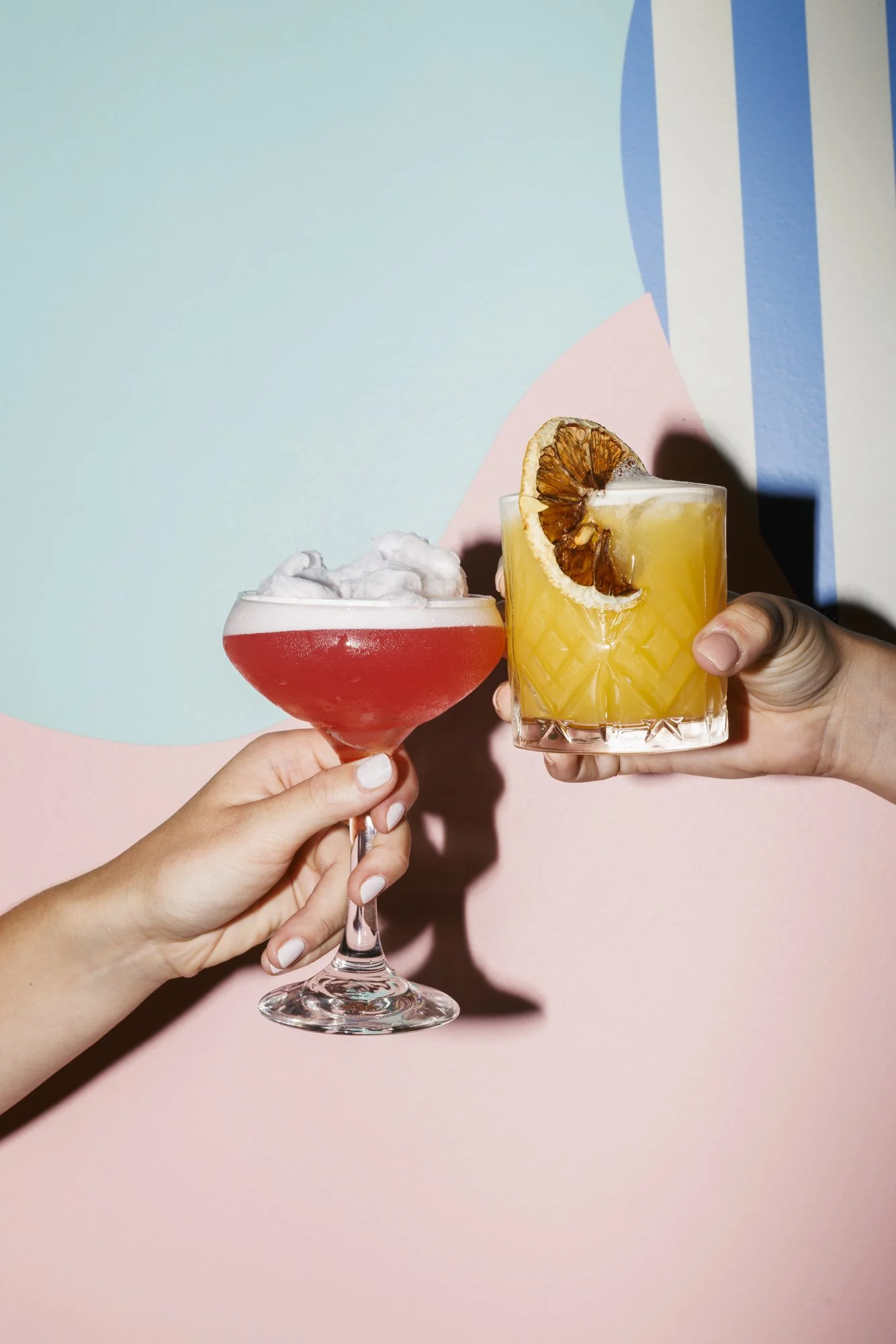Two hands holding different cocktails in front of a colorful, abstract background. One cocktail is a red drink with foam, served in a coupe glass. The other is a yellow drink with a dried citrus slice garnish, served in a short tumbler.