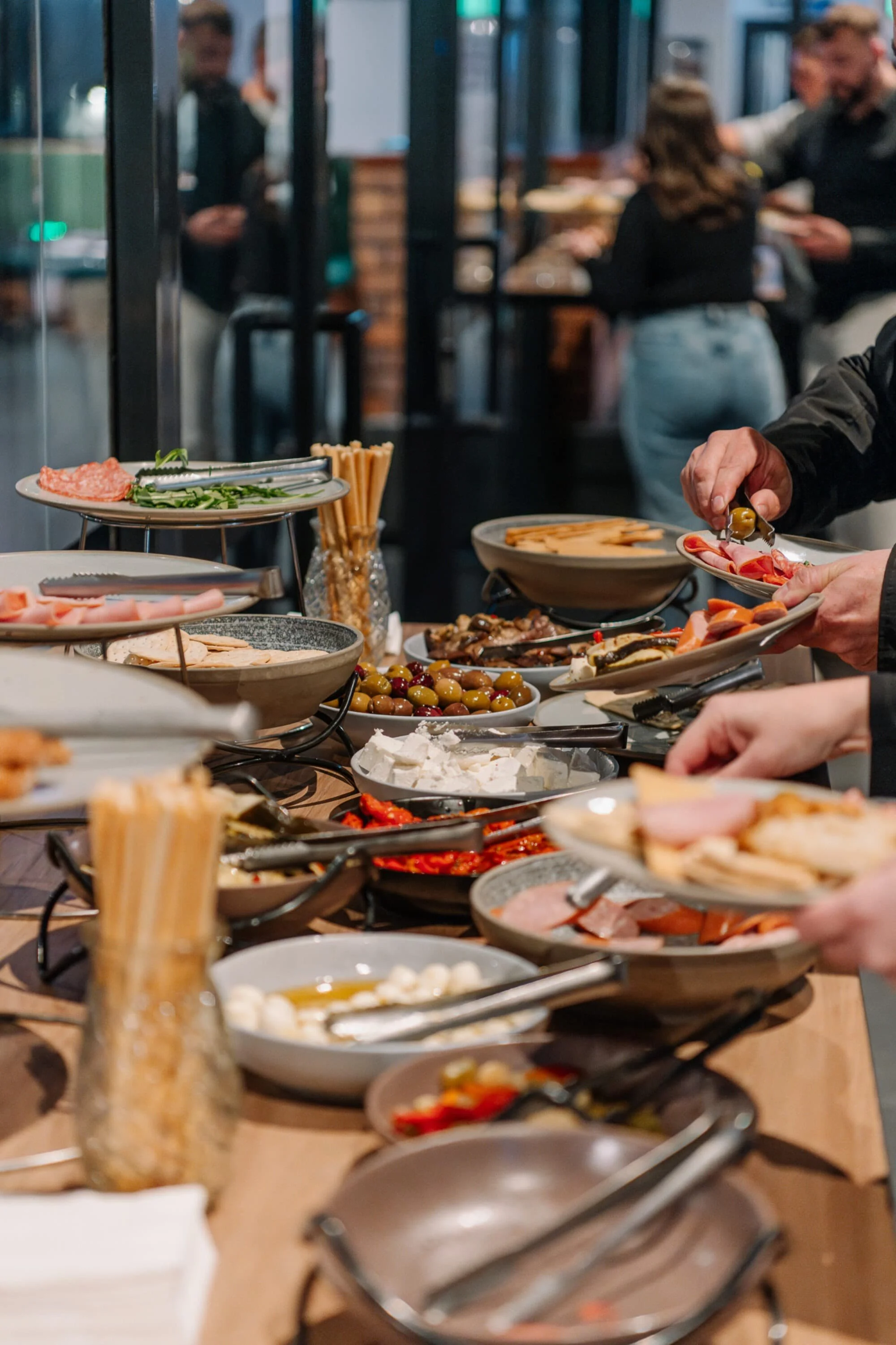 A buffet table with various appetizers including olives, cheeses, cured meats, breadsticks, and vegetables, with people serving themselves in the background.