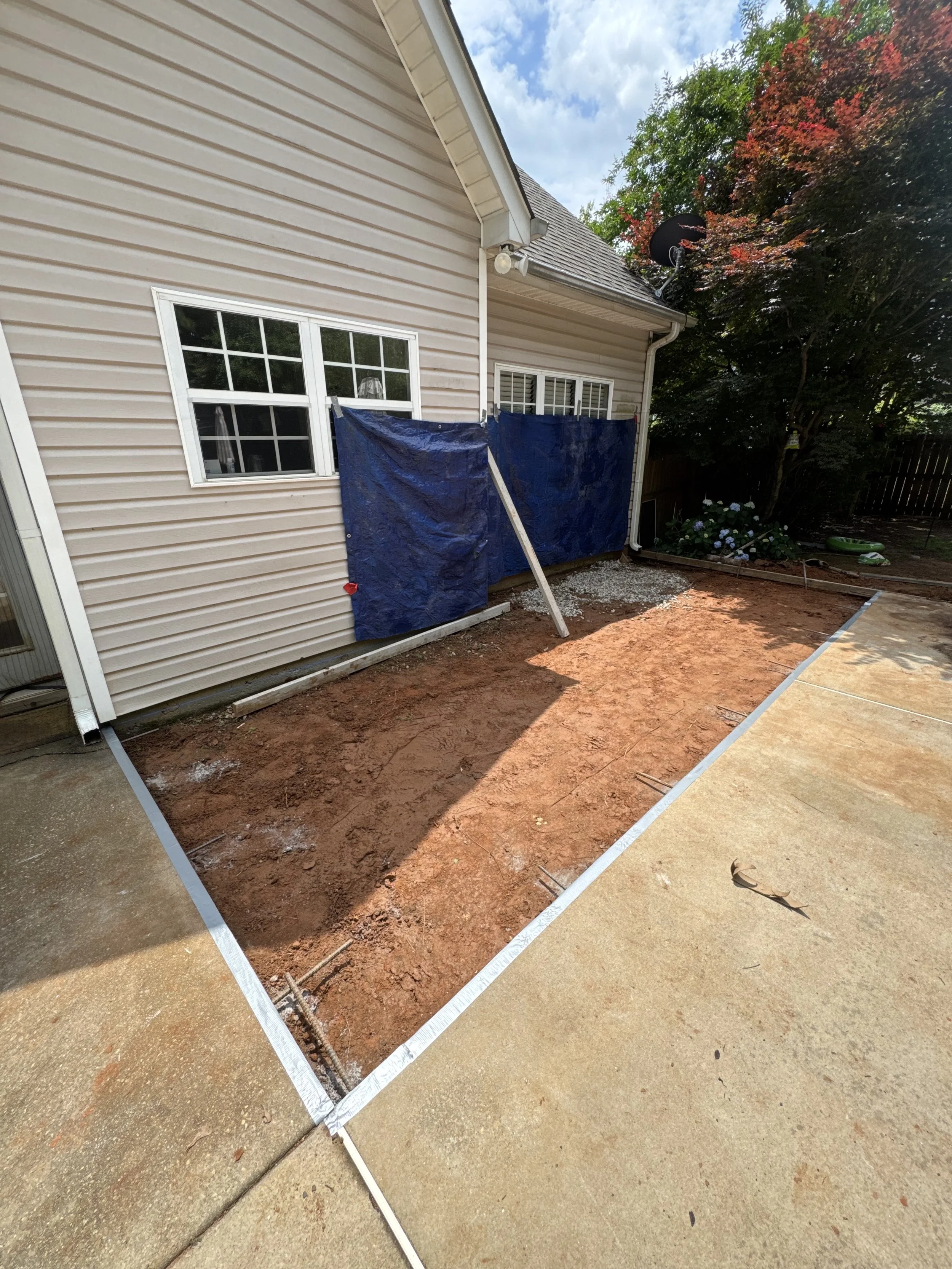 Backyard area under construction with exposed dirt, adjacent to a house with beige siding, featuring a window with white trim, and a blue tarp covering part of the backyard, with trees and a partly cloudy sky in the background.