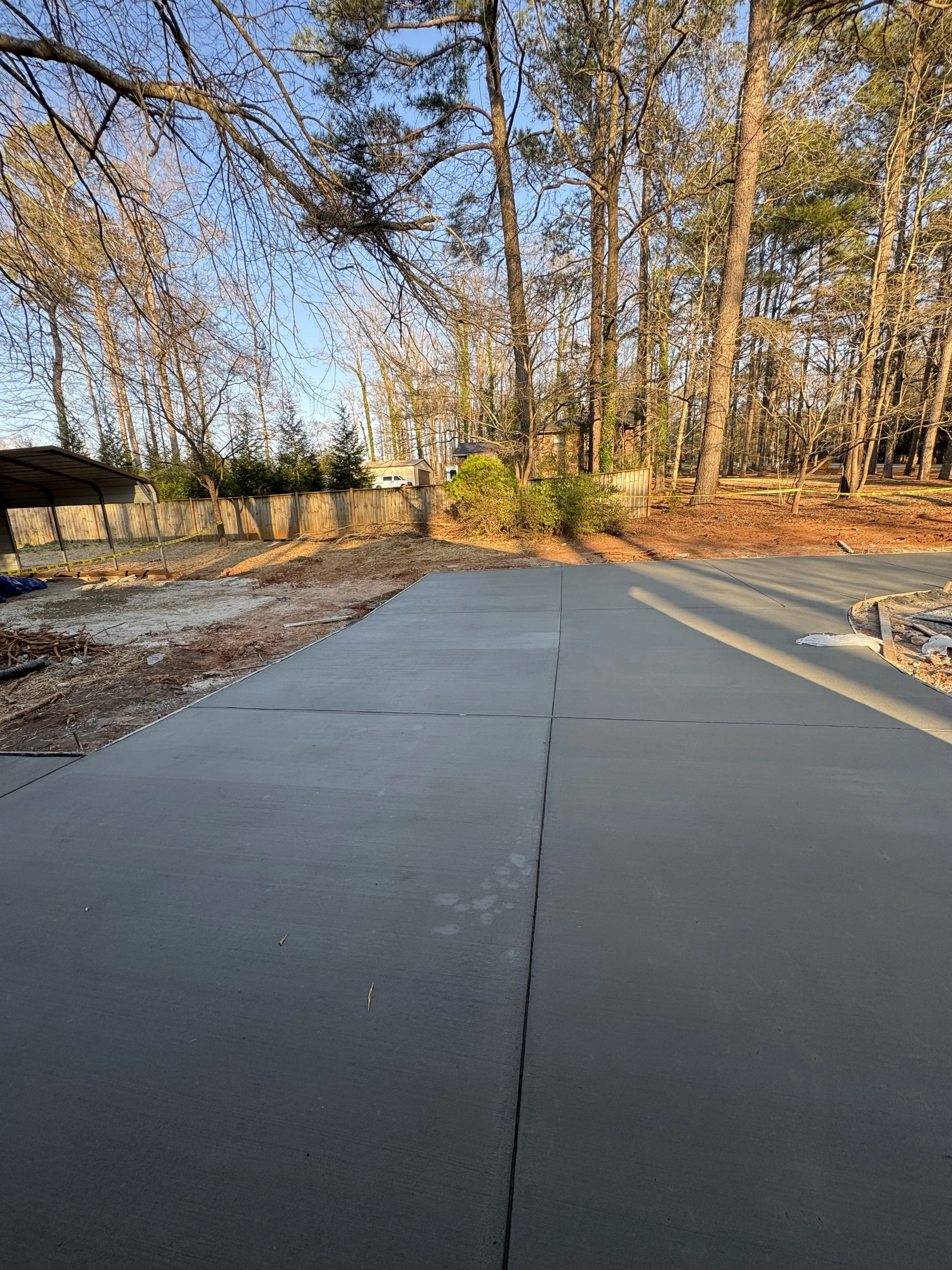 Freshly poured concrete patio with a view of trees and a backyard, sunset lighting, and a fence in the background.