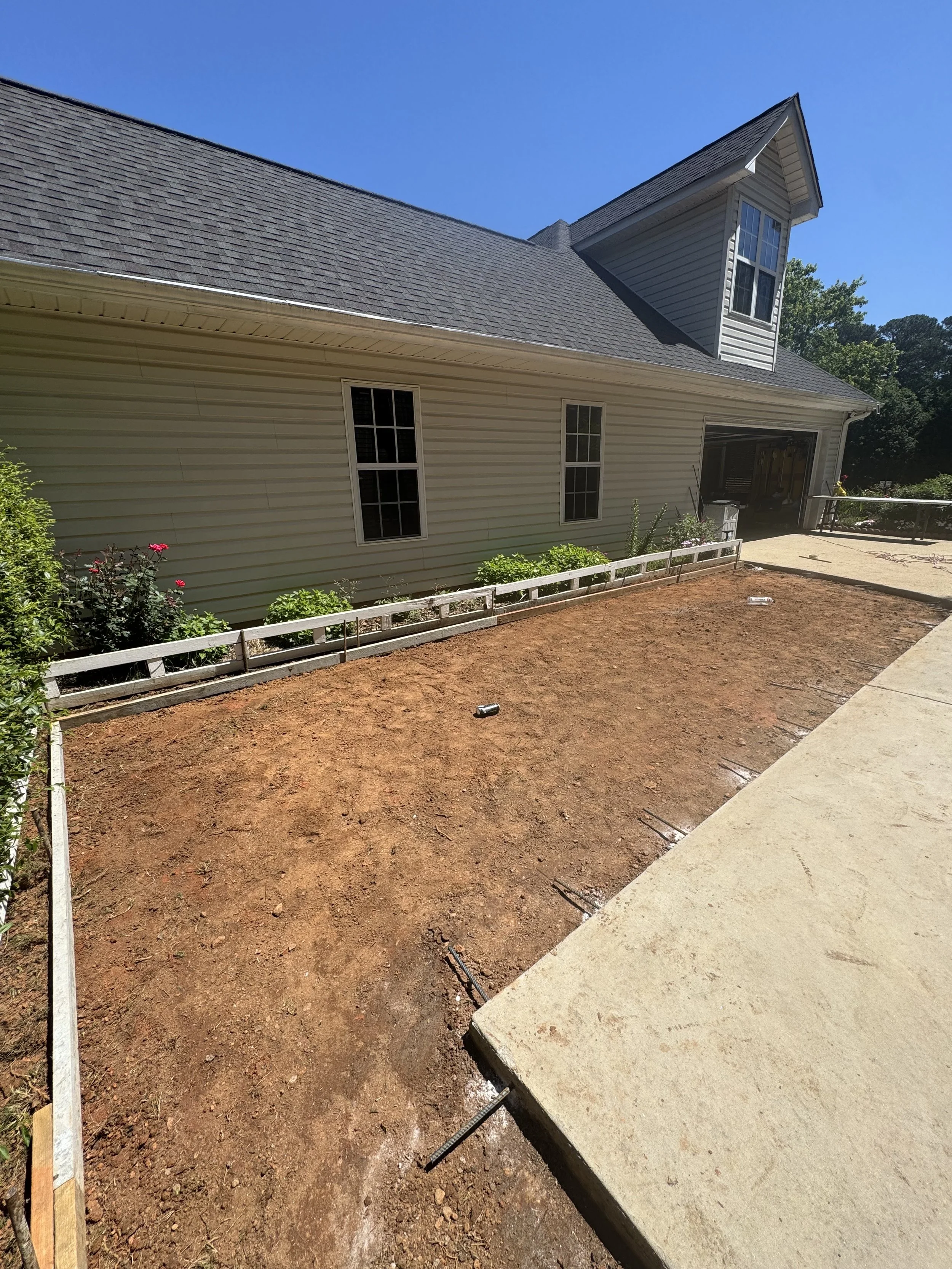A backyard with a recently paved concrete patio area and bare dirt ground, surrounded by a garden bed with plants, next to a house with beige siding and a gray shingle roof, under a blue sky.