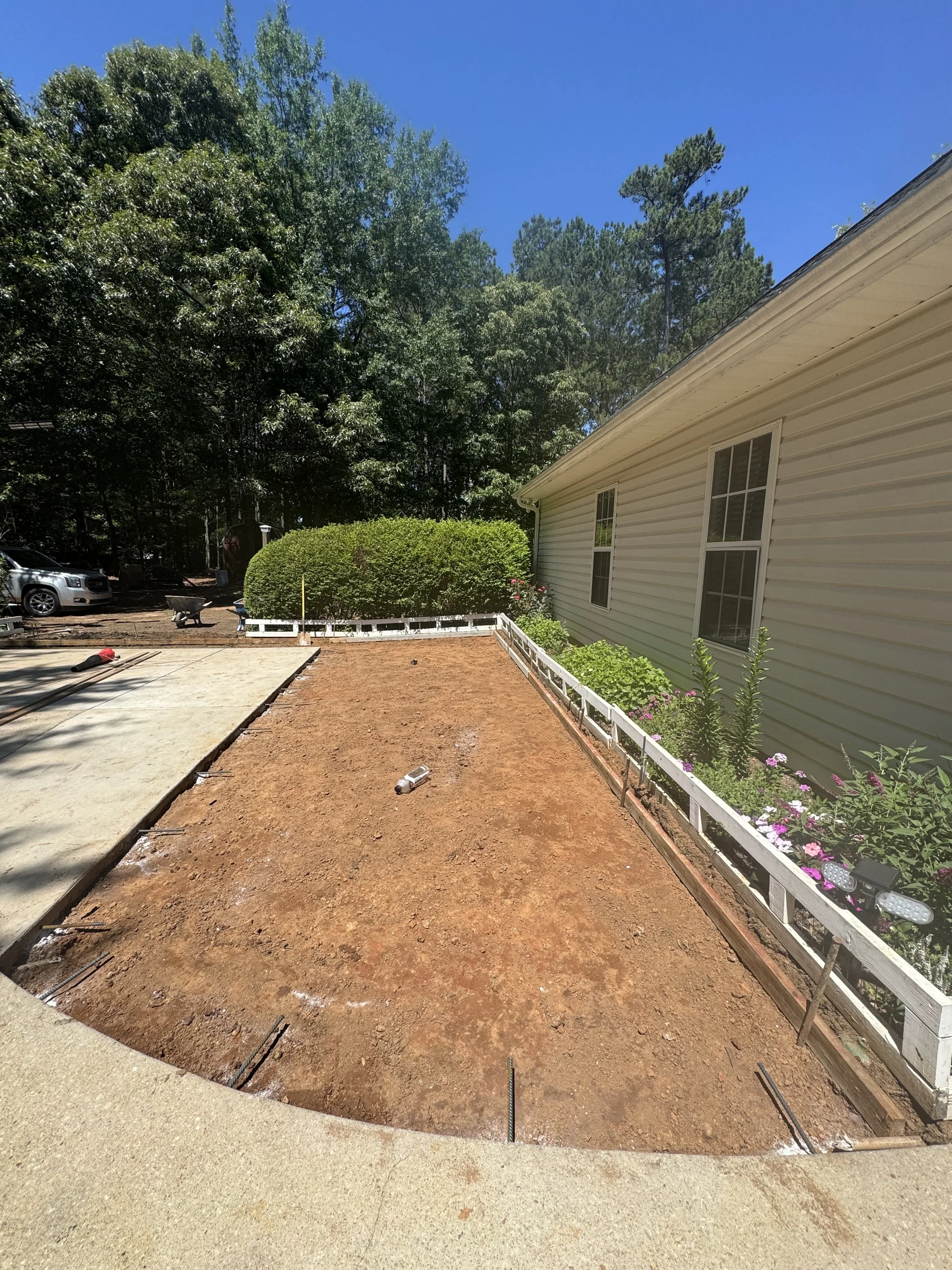 Lawn area in front of house under construction, with dirt and some construction tools, next to flower bed with plants and flowers along the house, and a backdrop of trees and a clear blue sky.