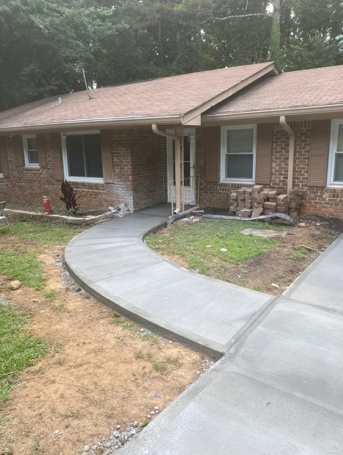 Newly poured concrete sidewalk in front of a brick house with brown shutters and a small garden bed.