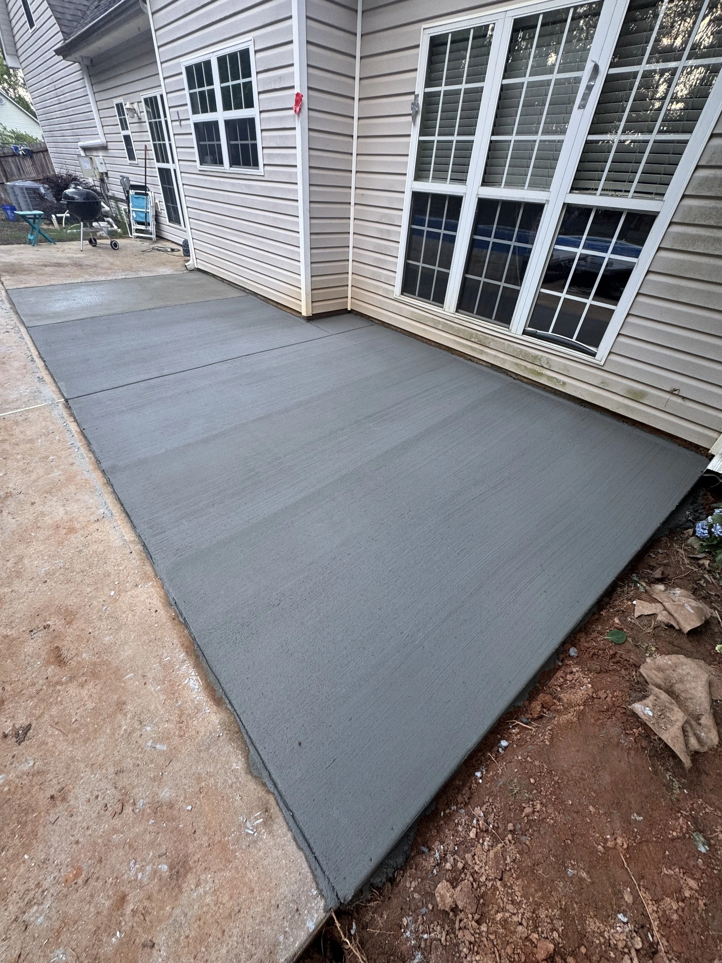 Recently poured concrete patio adjacent to a beige house with white window frames and shutters.