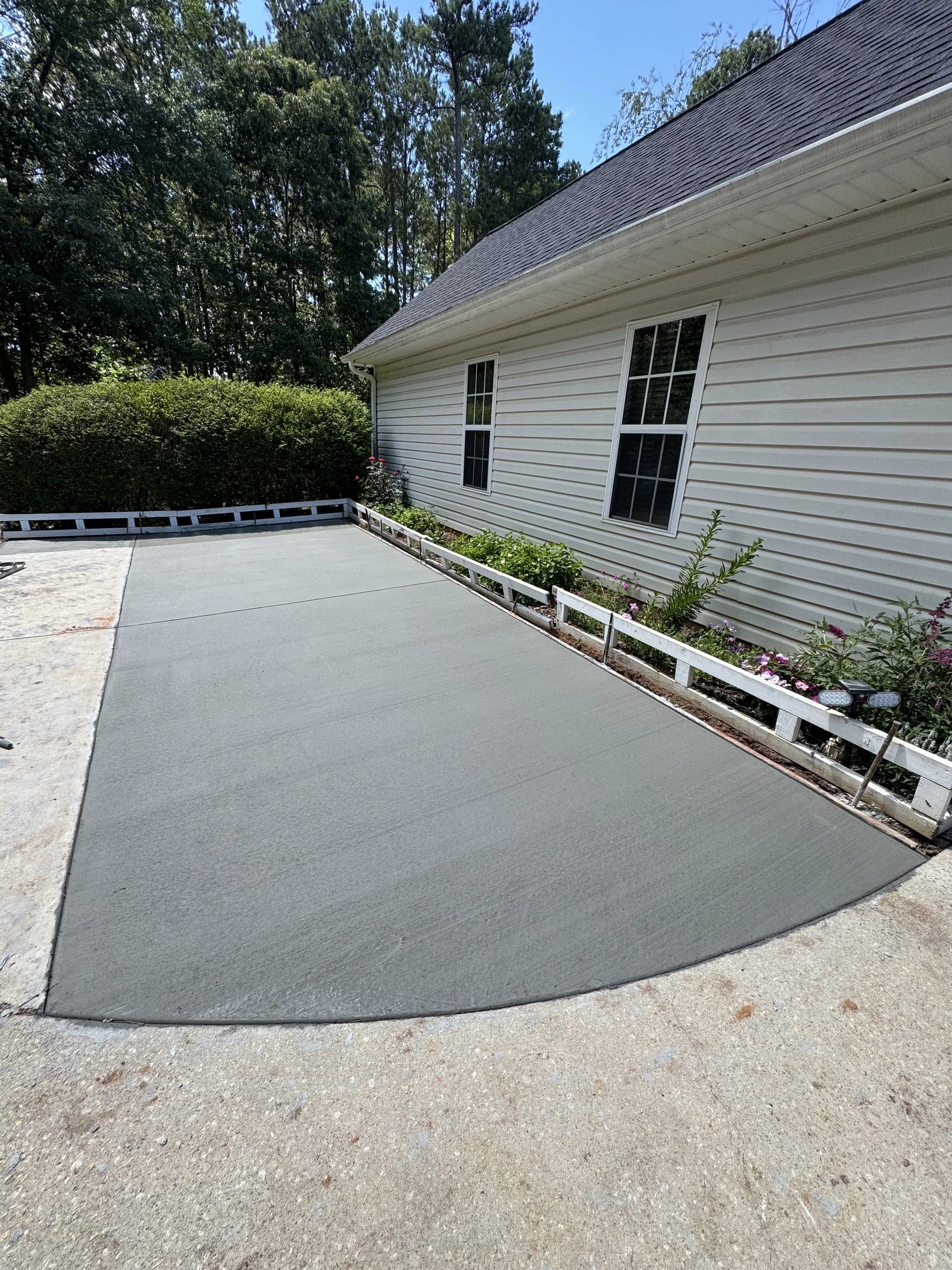 Smooth concrete driveway next to a beige house with white trim and two windows, bordered by flower beds with bushes and flowers, and surrounded by trees.