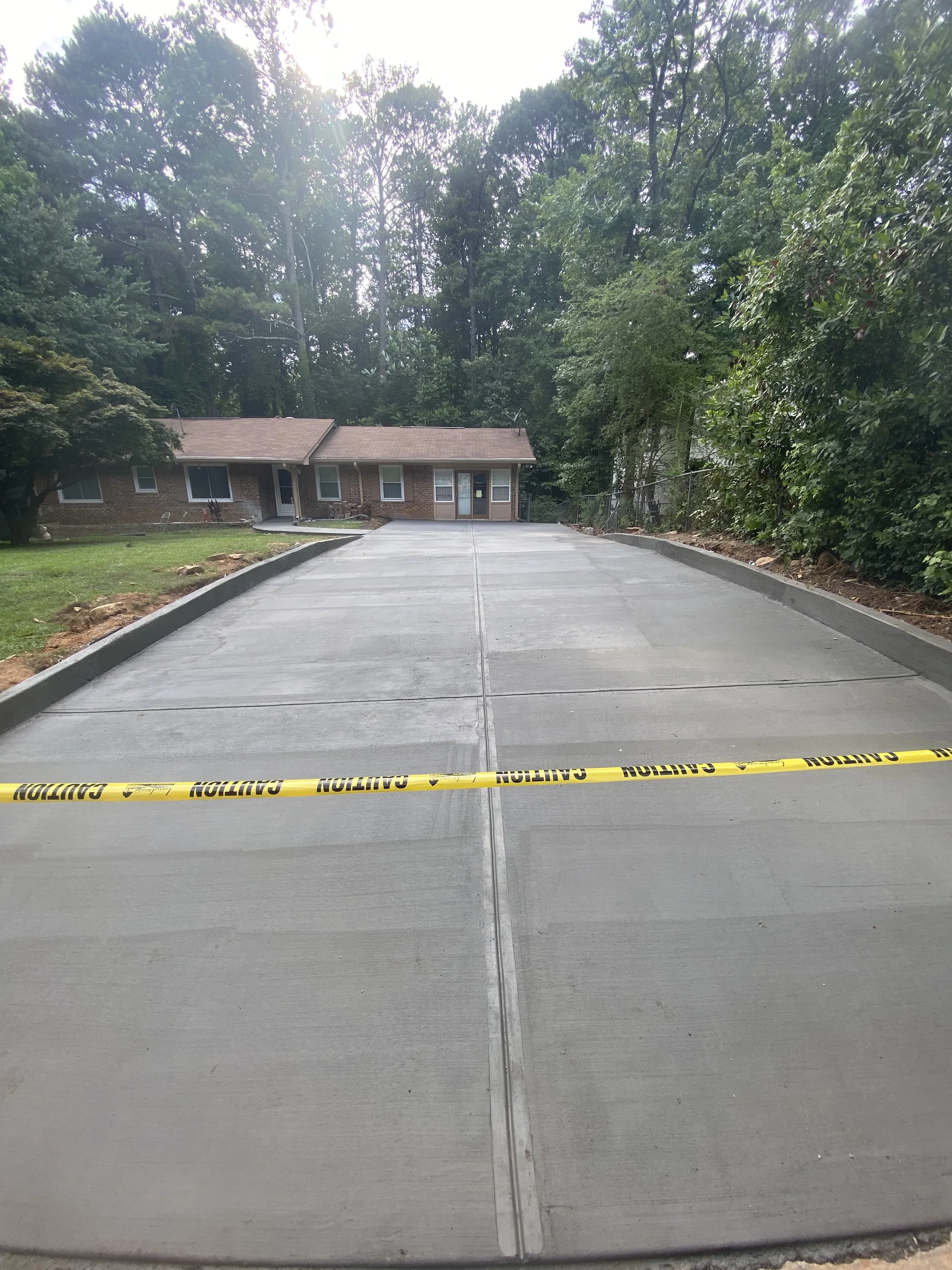 A freshly paved concrete driveway with yellow caution tape across the front, leading to a brick house with a brown roof and surrounded by trees.