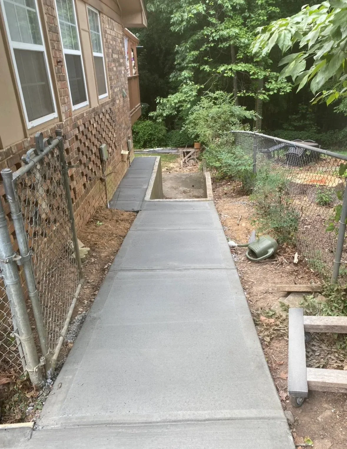 Newly poured concrete sidewalk next to a brick house, with a chain-link fence on the right, and lush green trees in the background.