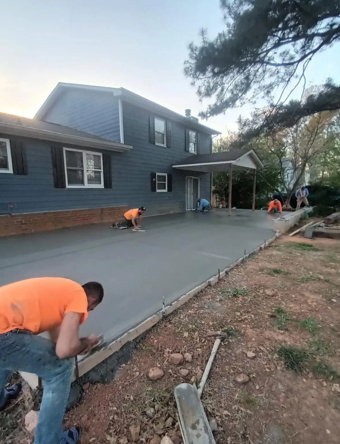 Workers in orange shirts are smoothing wet concrete to create a new patio or walkway in front of a blue house, with construction tools and dirt surrounding the area.