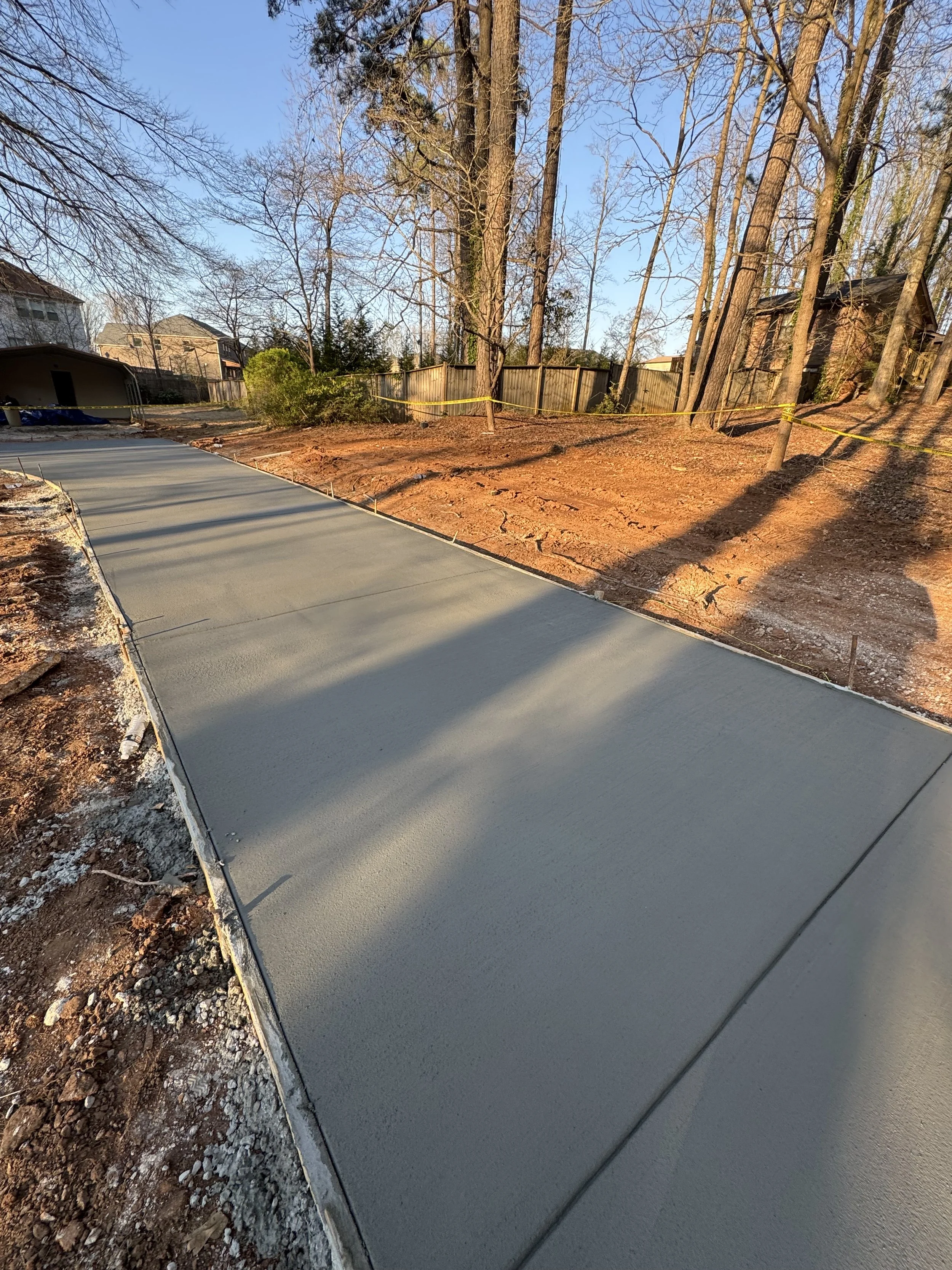 Freshly poured concrete sidewalk in a residential backyard, surrounded by dirt and trees, with a wooden fence in the background during late afternoon sunlight.