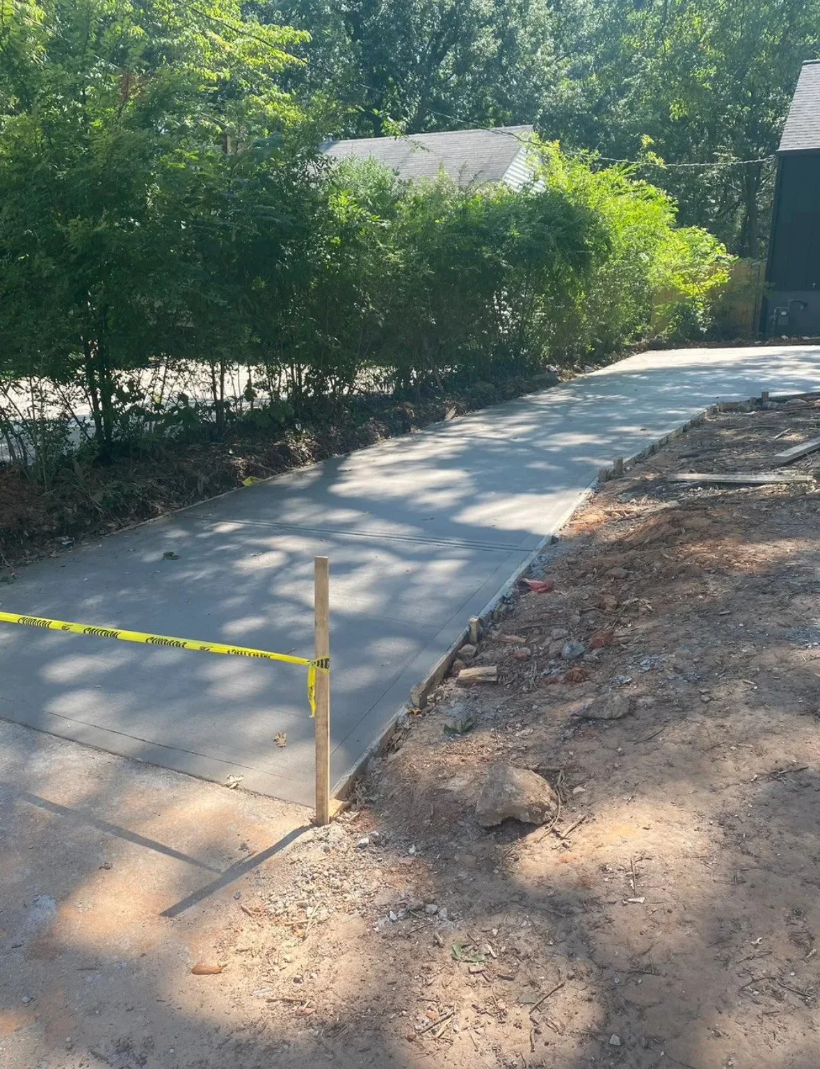 Newly poured concrete sidewalk under construction with wooden stakes and yellow caution tape, surrounded by dirt and greenery.