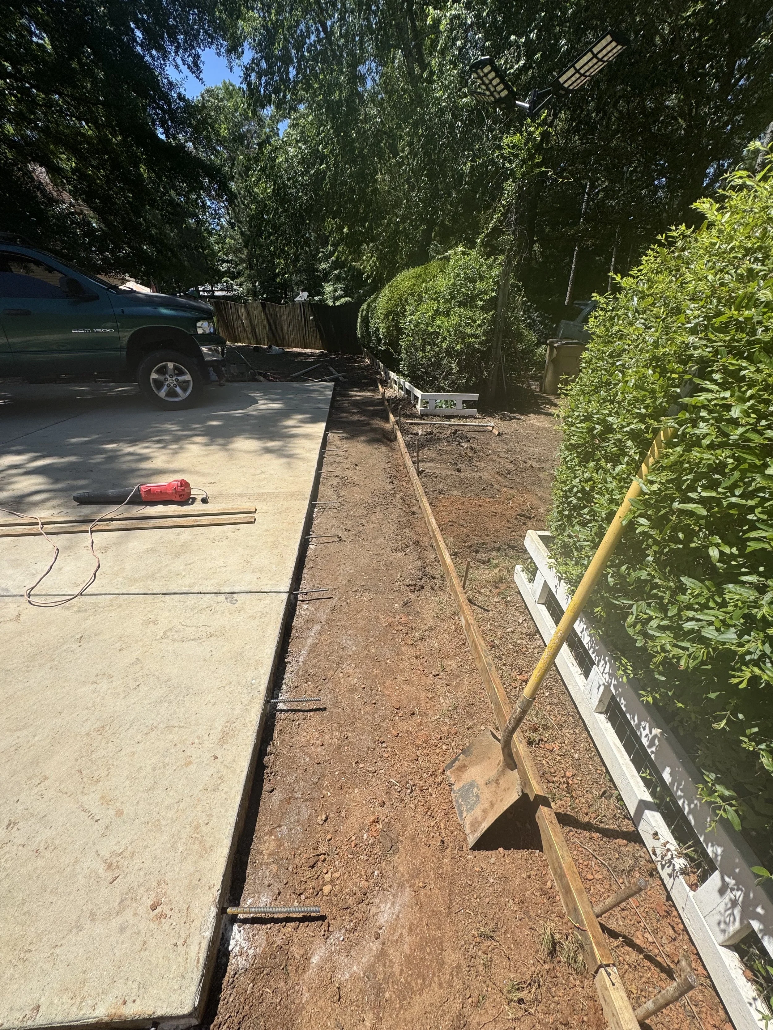 Construction site showing a concrete driveway, a parked truck, a shovel, and a garden bed being prepared for planting.