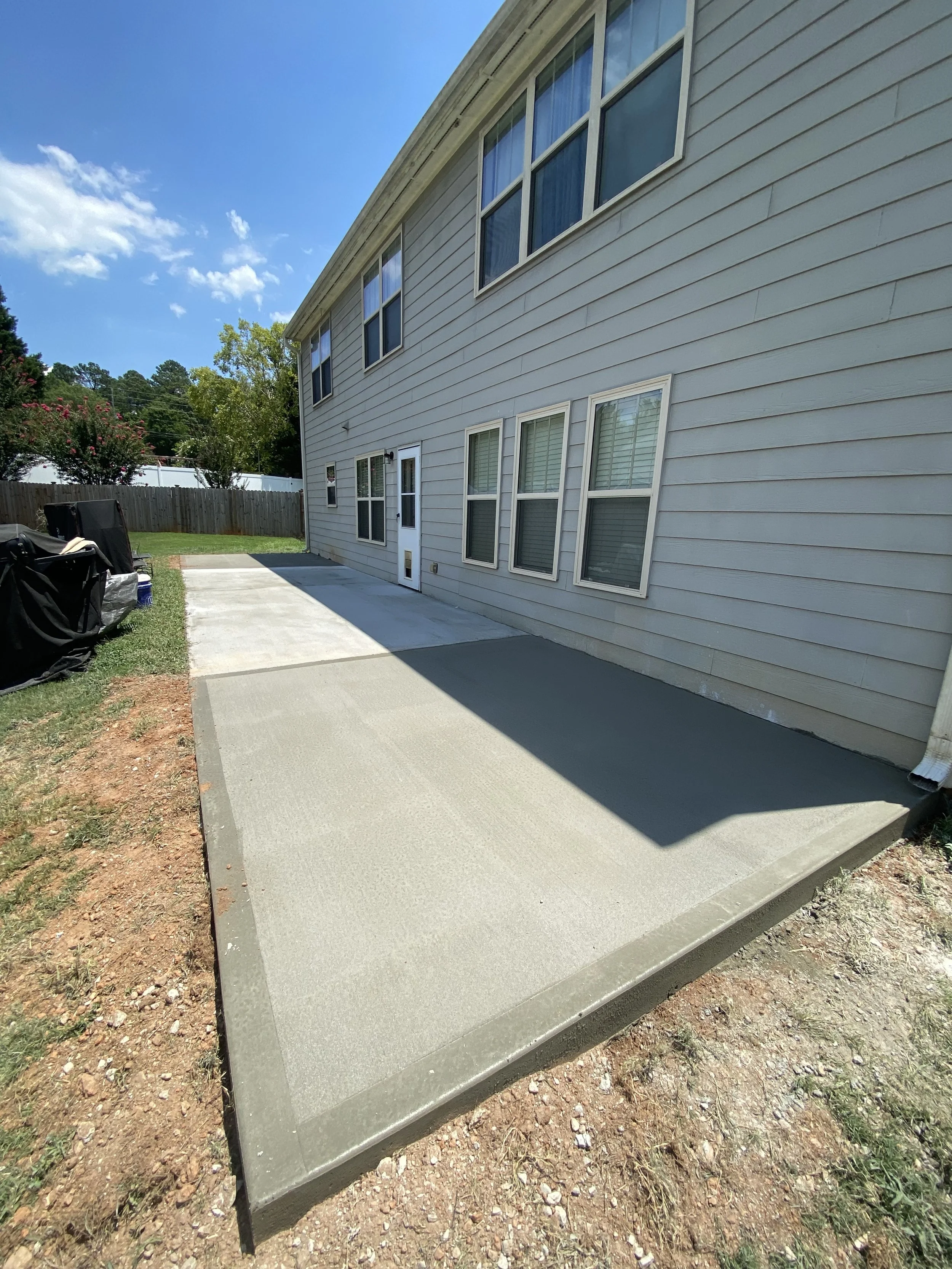 Newly poured concrete patio in the backyard of a house, with multiple windows and a side door, bordered by a wooden fence and some landscaping area.