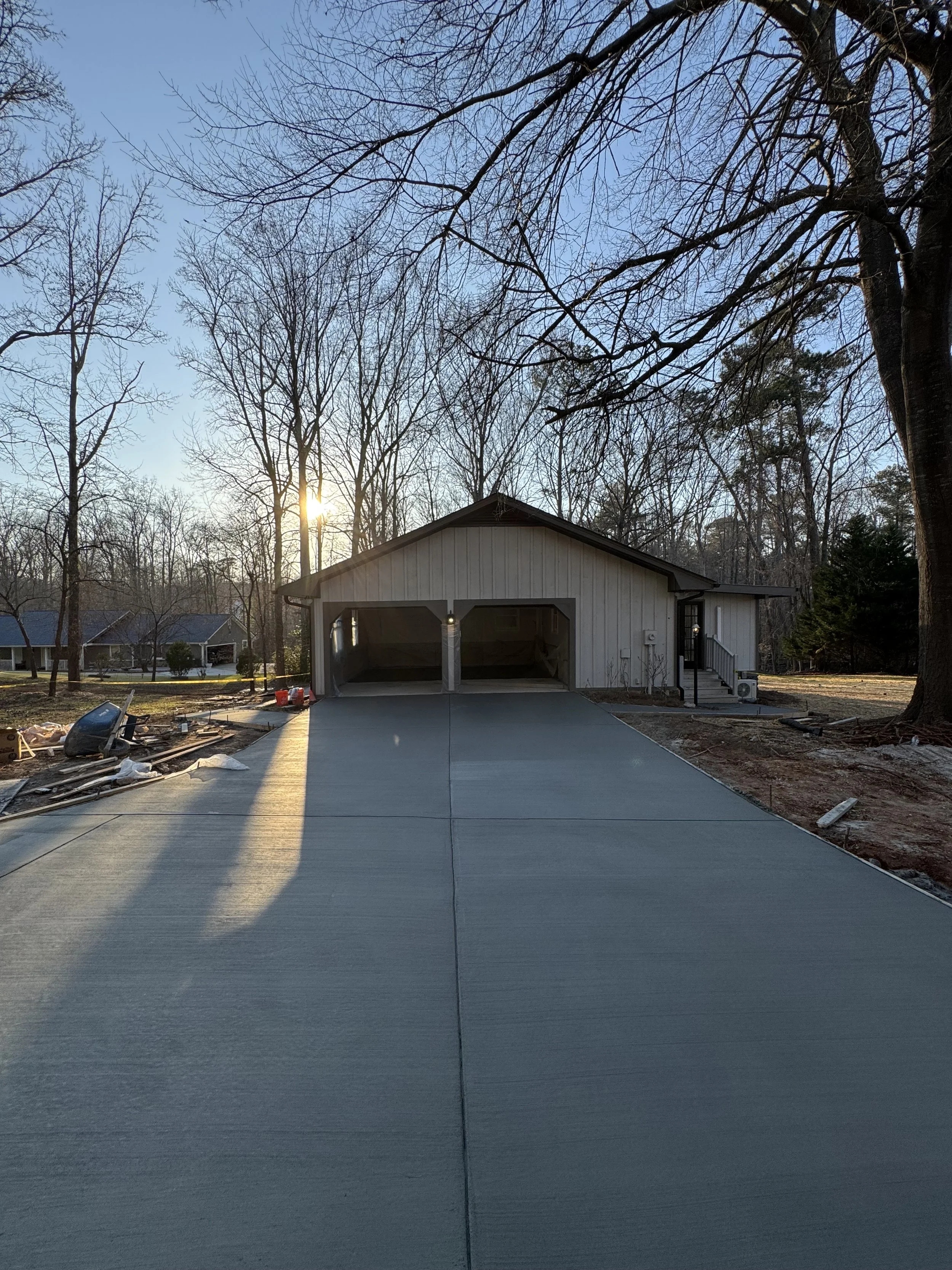 Newly constructed garage with open doors, concrete driveway leading up to it, trees in the background, and the sunset behind the trees.