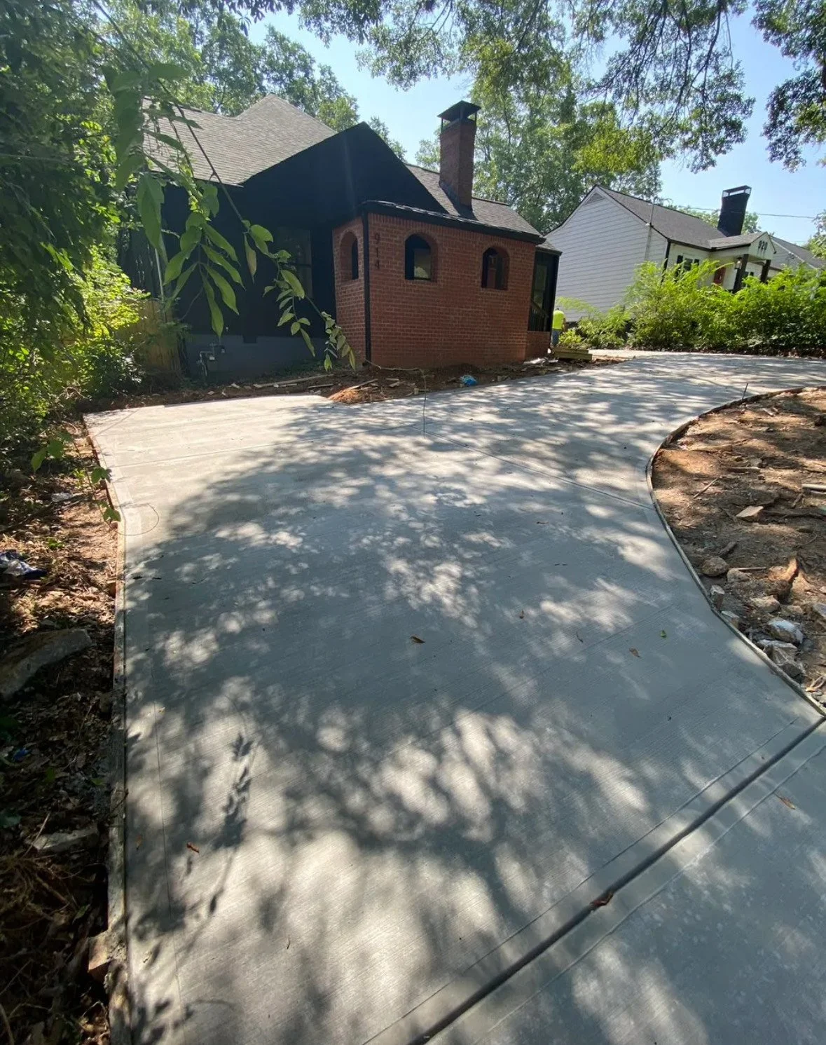 Newly poured concrete driveway in front of a brick house with chimney, surrounded by trees and other houses in a residential neighborhood.
