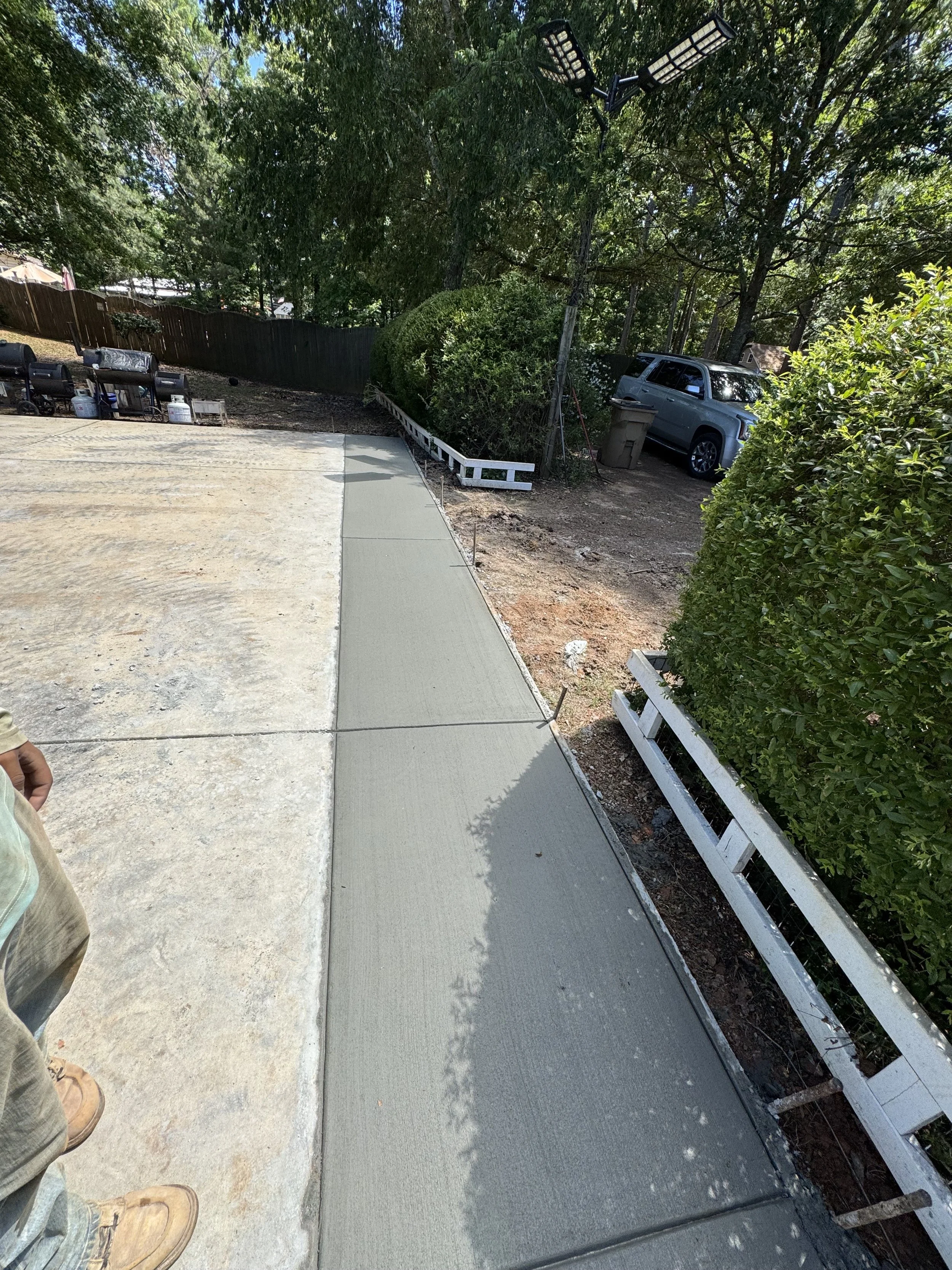 Newly poured concrete sidewalk next to an existing concrete slab, with construction tools, a person wearing work boots, and a white railing along the edge, with trees and a parked SUV in the background.