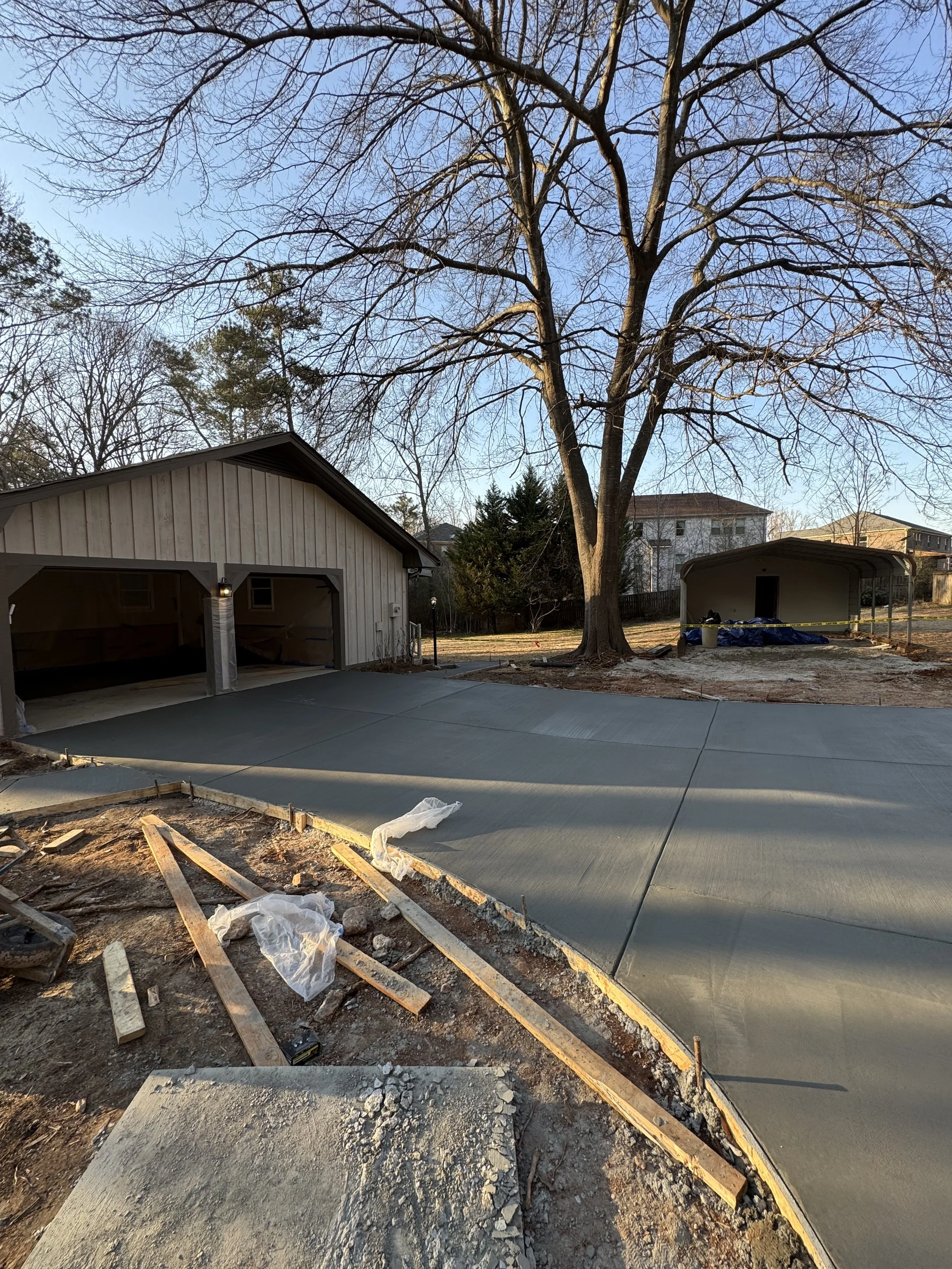 Concrete driveway under construction with wooden formwork and construction materials in front of a garage, with a large leafless tree and other houses in the background.