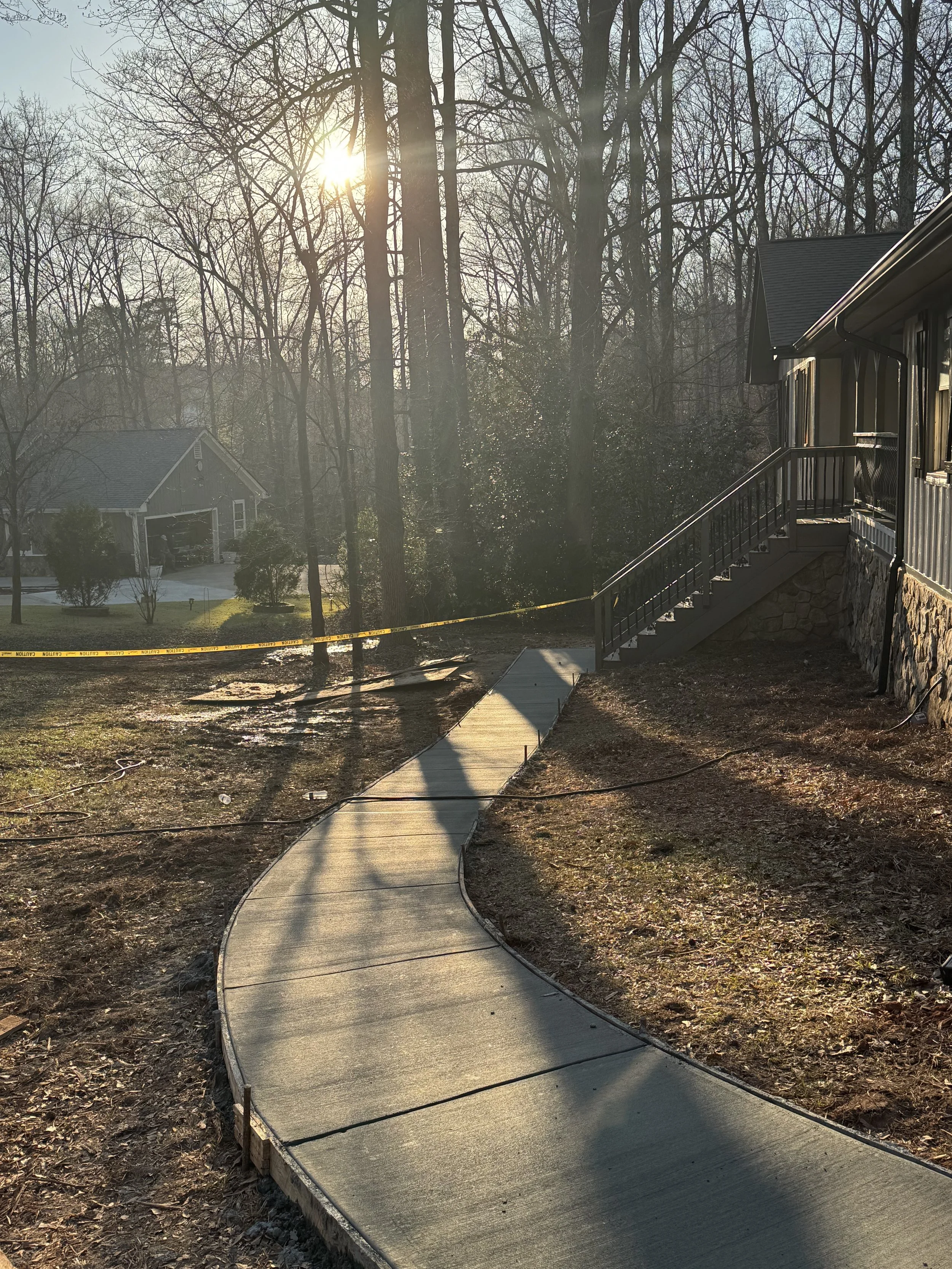 A curved concrete pathway leading to a house with wooden steps, trees in the background, and sunlight filtering through the trees, with a yellow caution tape across the yard.