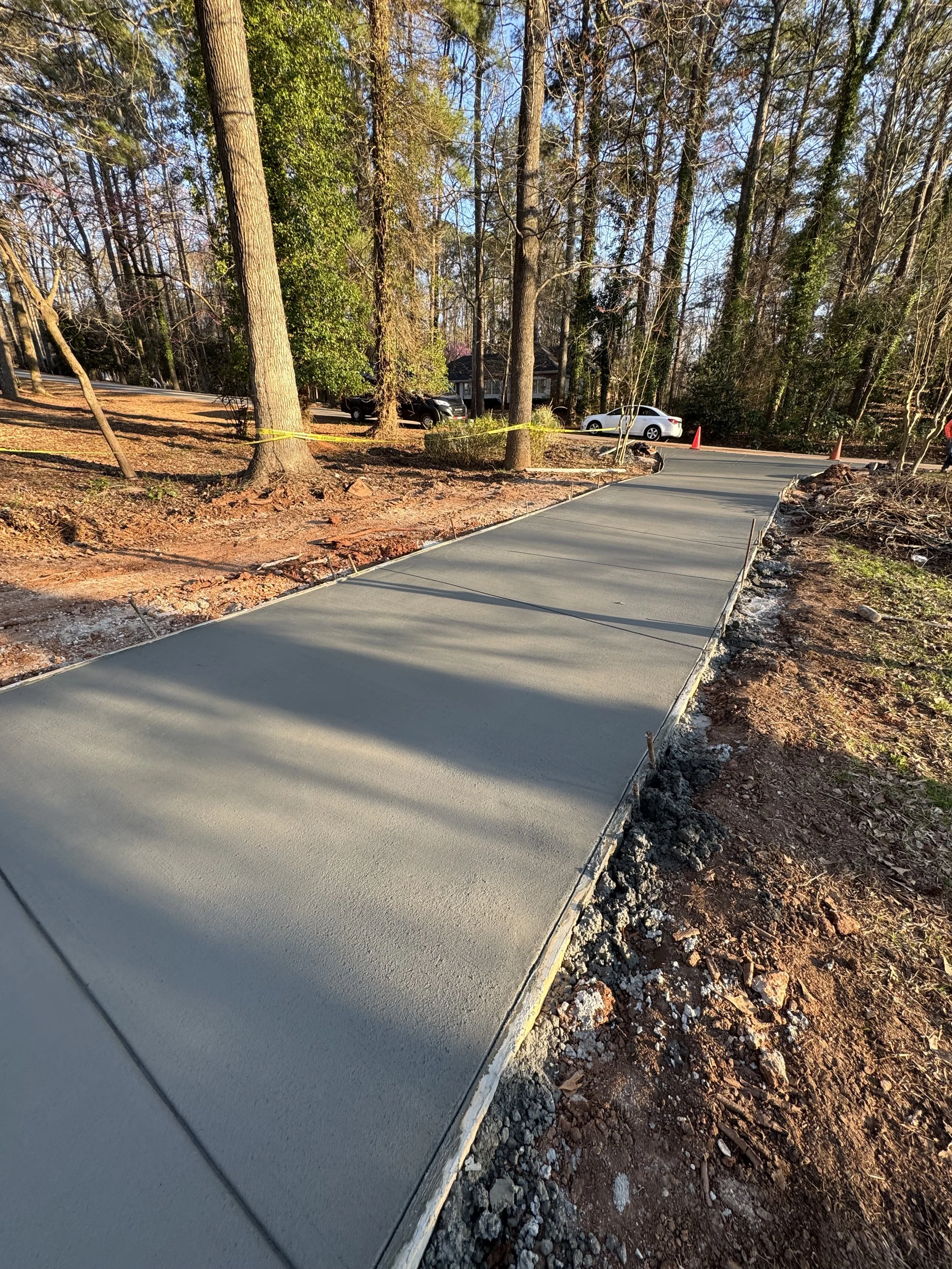Freshly poured concrete sidewalk under construction, with trees and parked cars in the background, and orange traffic cones