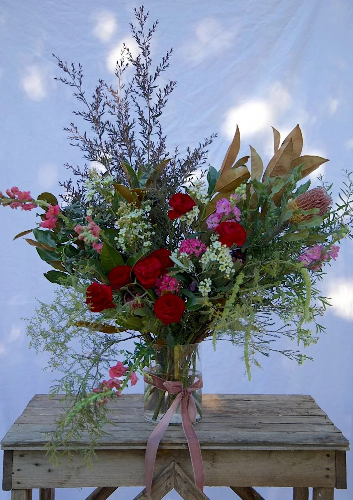 A large bouquet of mixed flowers in a glass vase with a pink ribbon, placed on a rustic wooden table, against a light blue sky with scattered clouds.