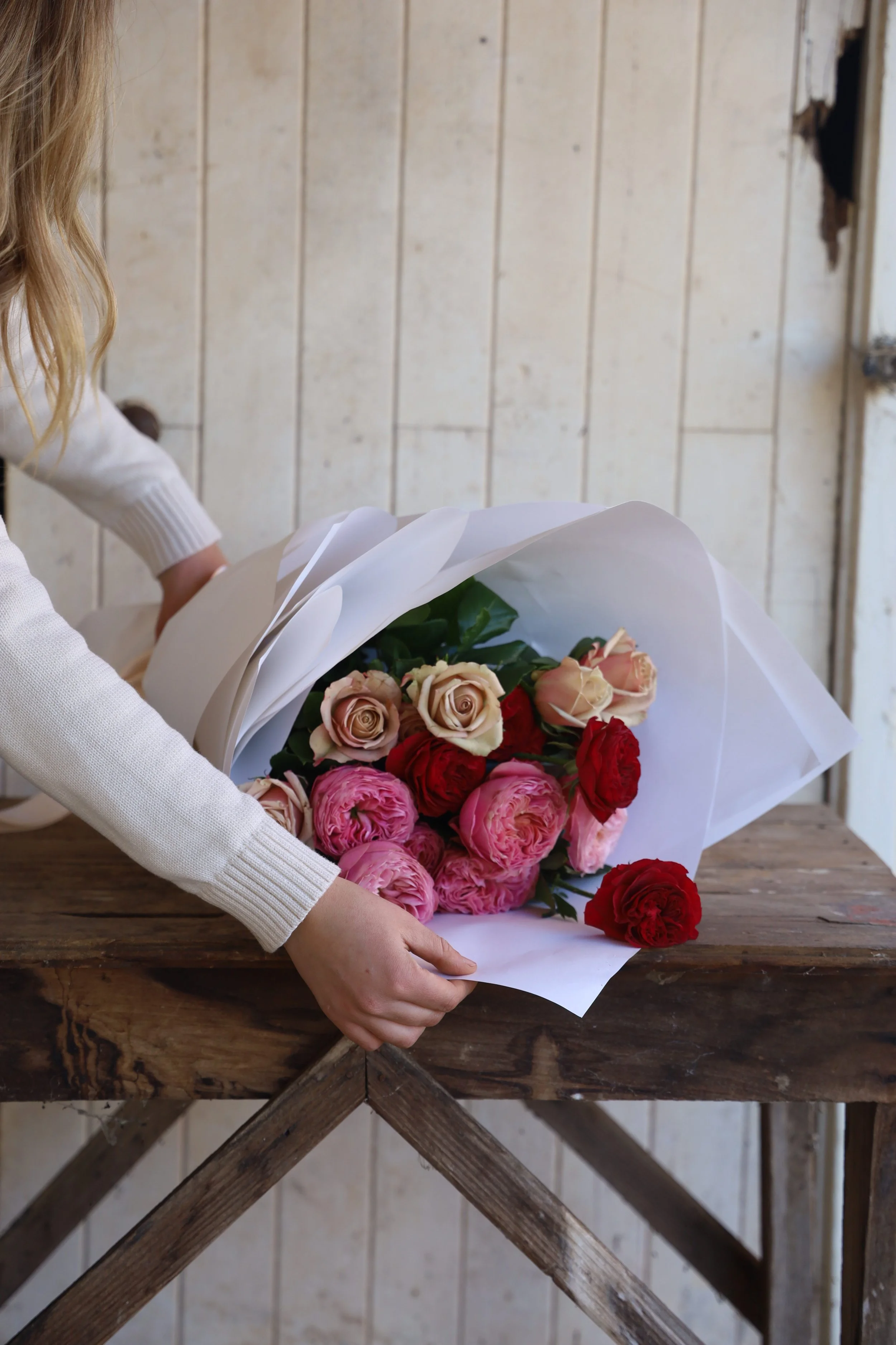 Person arranging a bouquet of pink, red, and cream roses on a wooden table in a rustic setting.