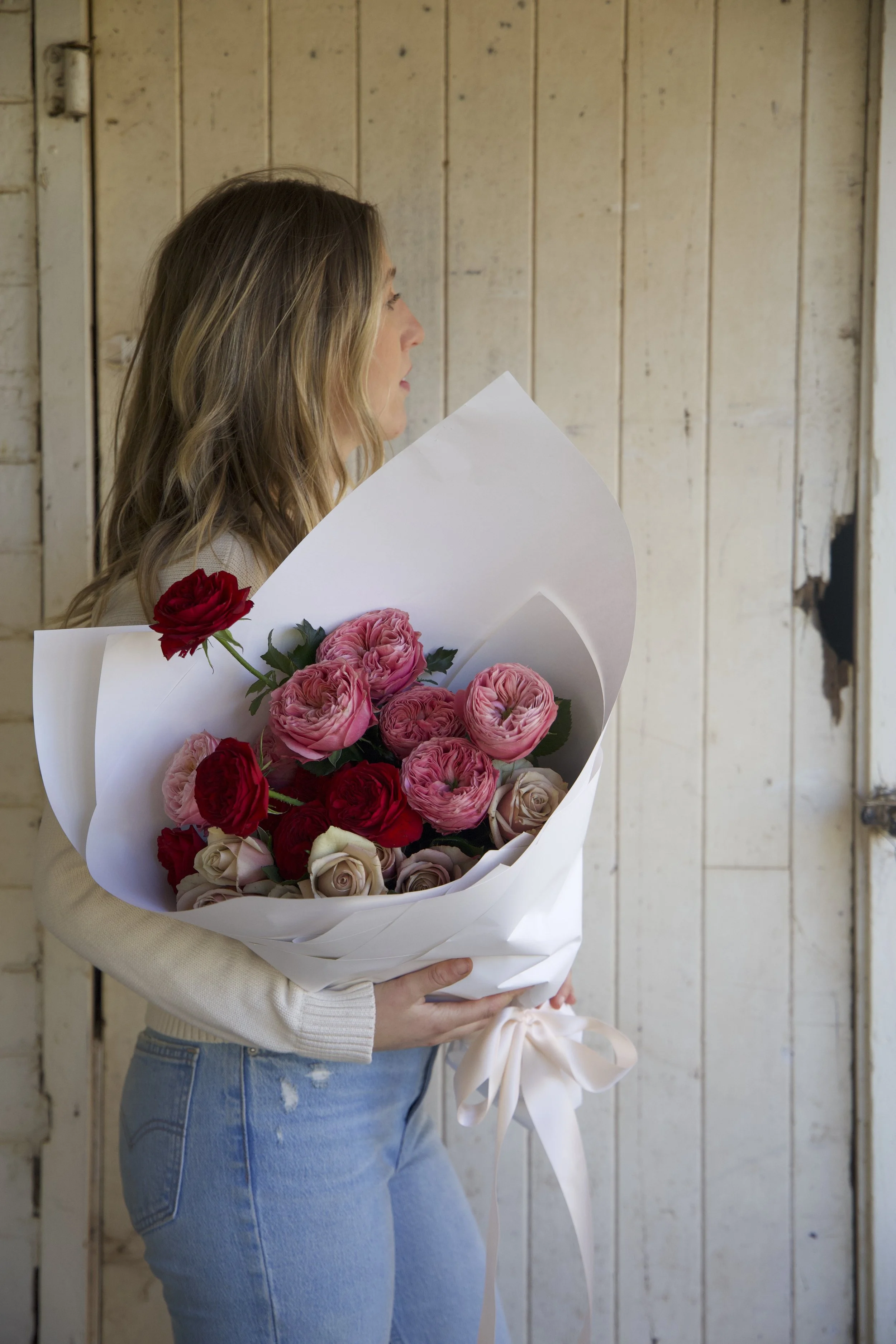 Woman holding a bouquet of pink, red, and cream roses in a white wrapping paper with a bow, standing in front of a rustic wooden wall.
