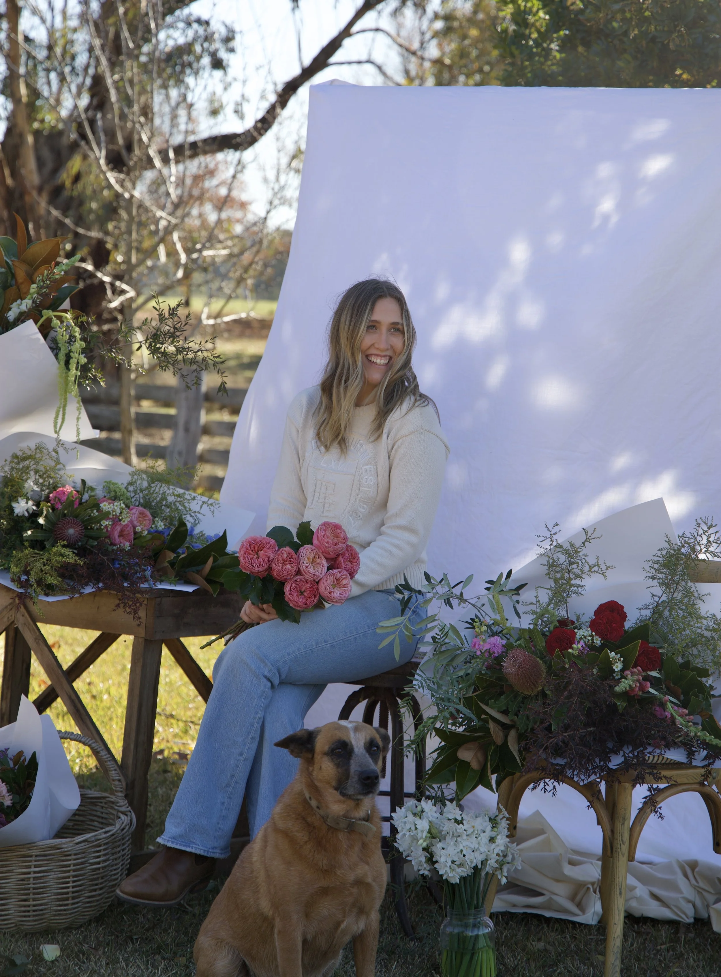 A woman sitting outdoors with a dog, surrounded by flowers and greenery, with a white backdrop behind her.