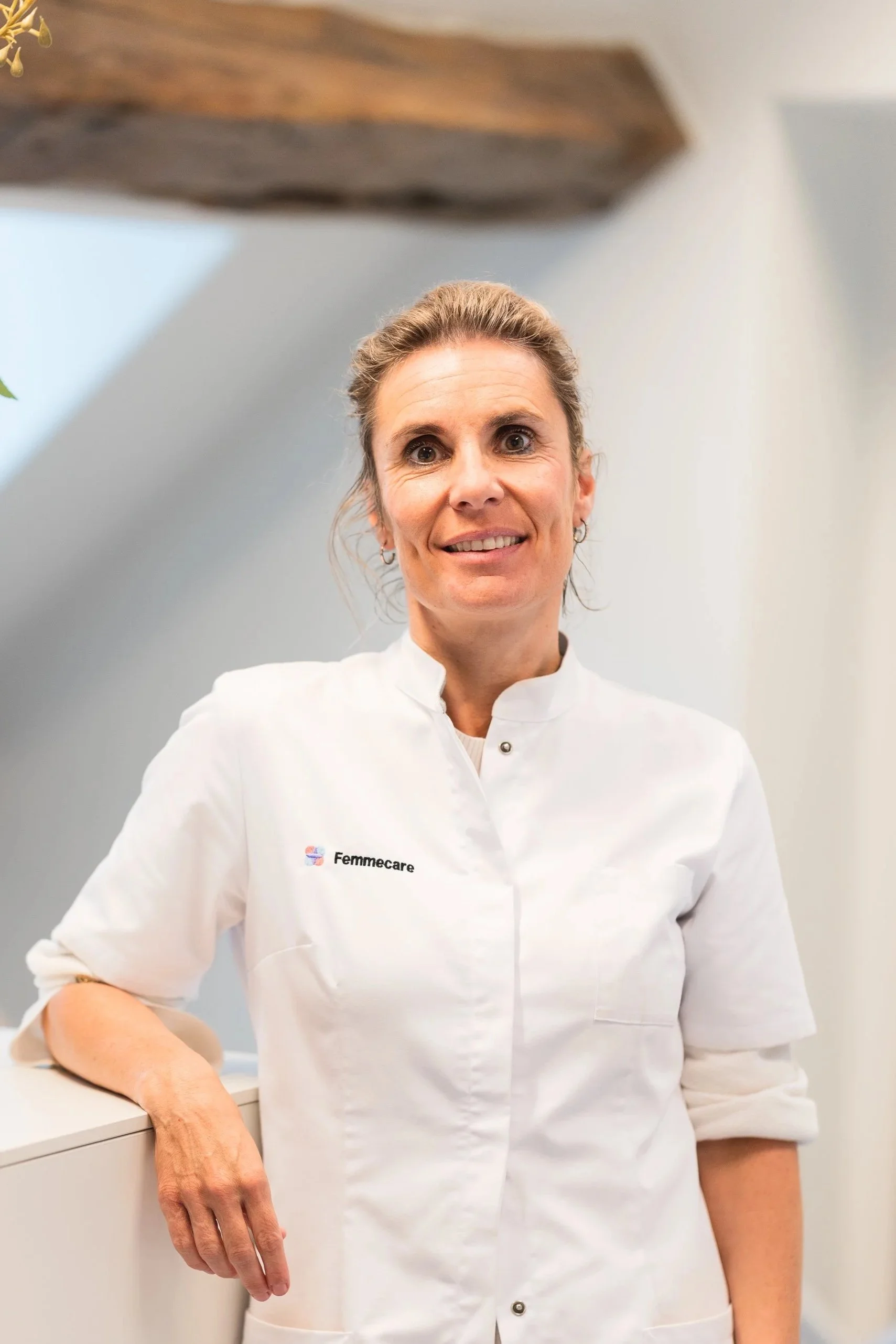 Close-up van een glimlachende vrouw in een witte verpleegstersjas met logo 'Femmcare', staand in een kliniek of medische omgeving.