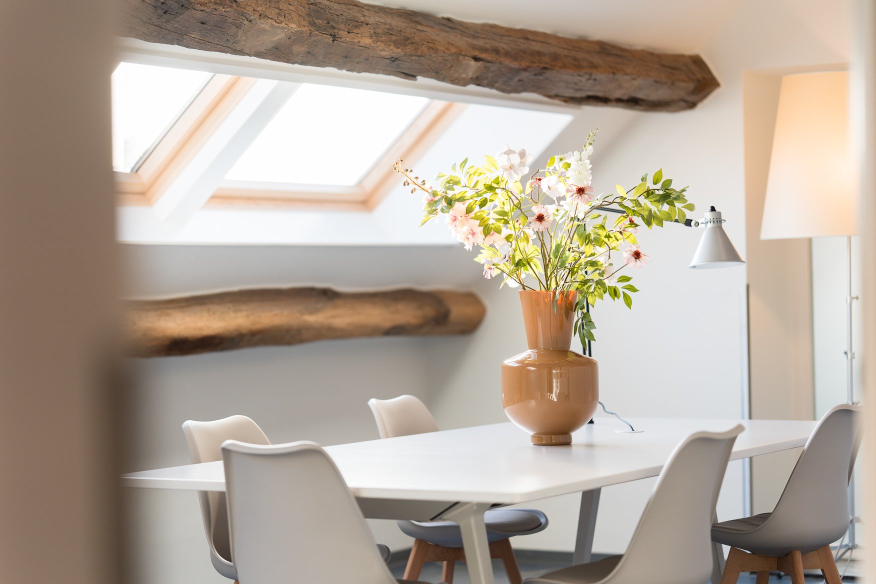 Een witte eetkamer tafel met een grote terracotta vaas met bloemen, onder een dakraam met houten balken op het plafond.