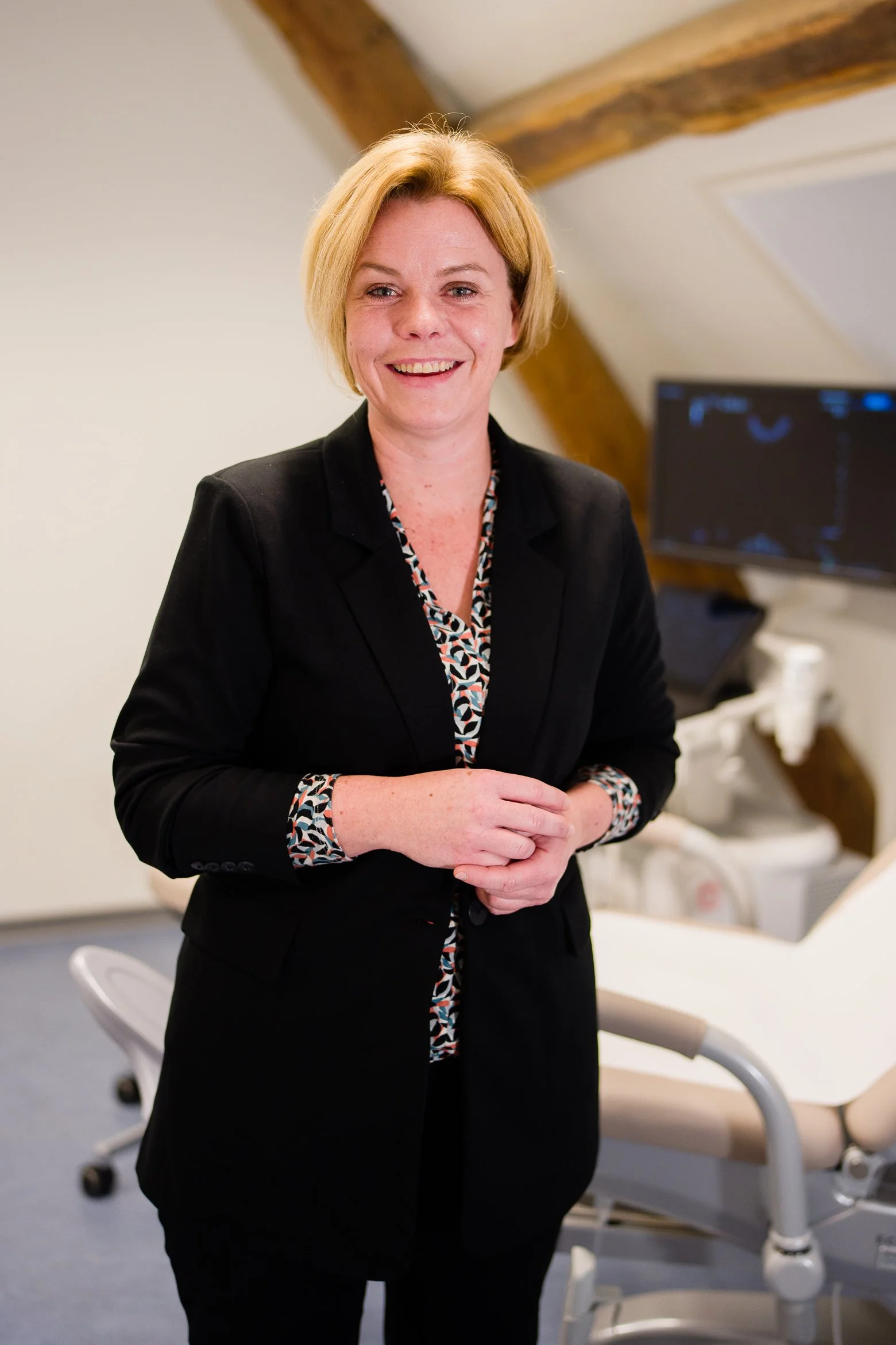 Een lachende vrouw in een zwarte outfit in een medische omgeving, met een doktersstoel en medische apparatuur op de achtergrond.