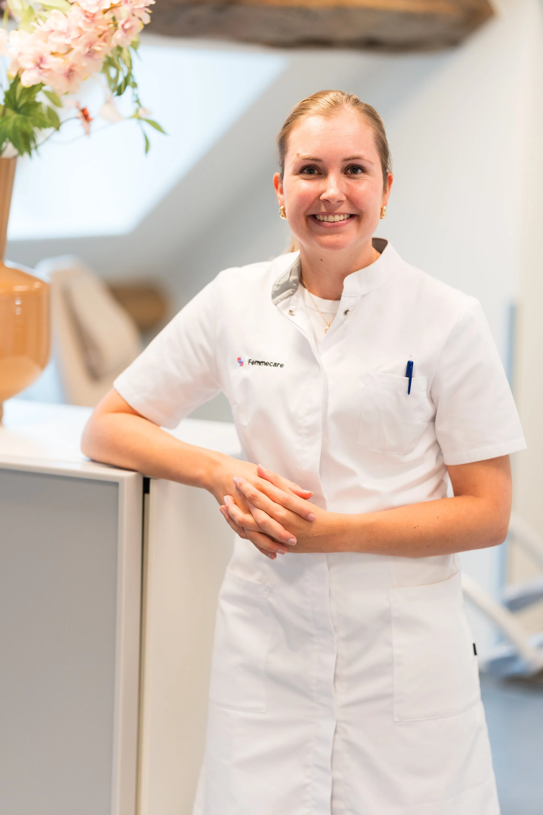 Een glimlachende vrouwelijke verpleegkundige in een witte uniform poseert binnen, met een vaas met bloemen op de achtergrond.