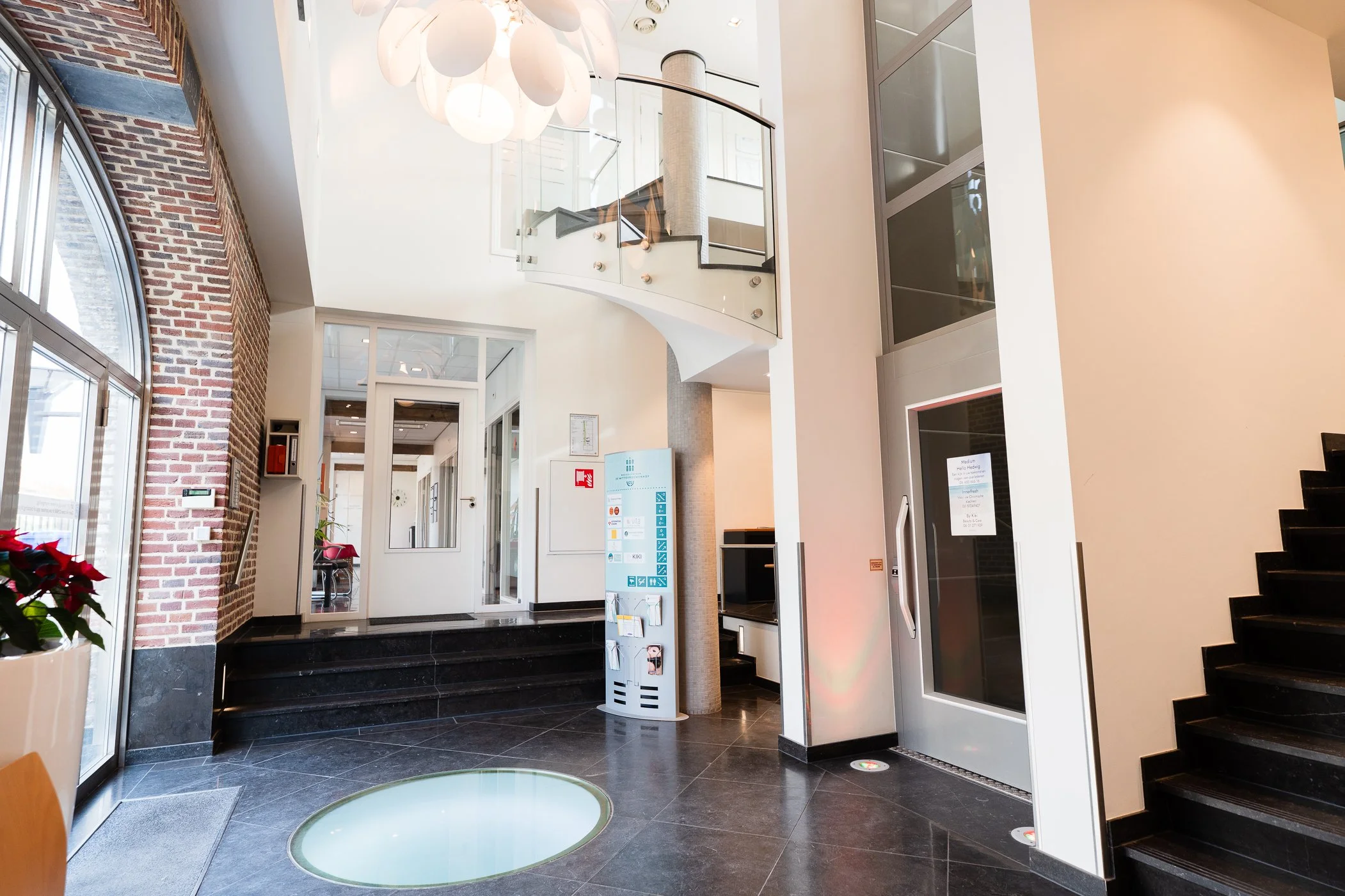 Interieur van een modern gebouw met zwarte tegelvloer, een glazen lift en een trap, grote ramen met boogvorm, een paddenstoelvormige plafondlamp, en een whiteboard op een muur.