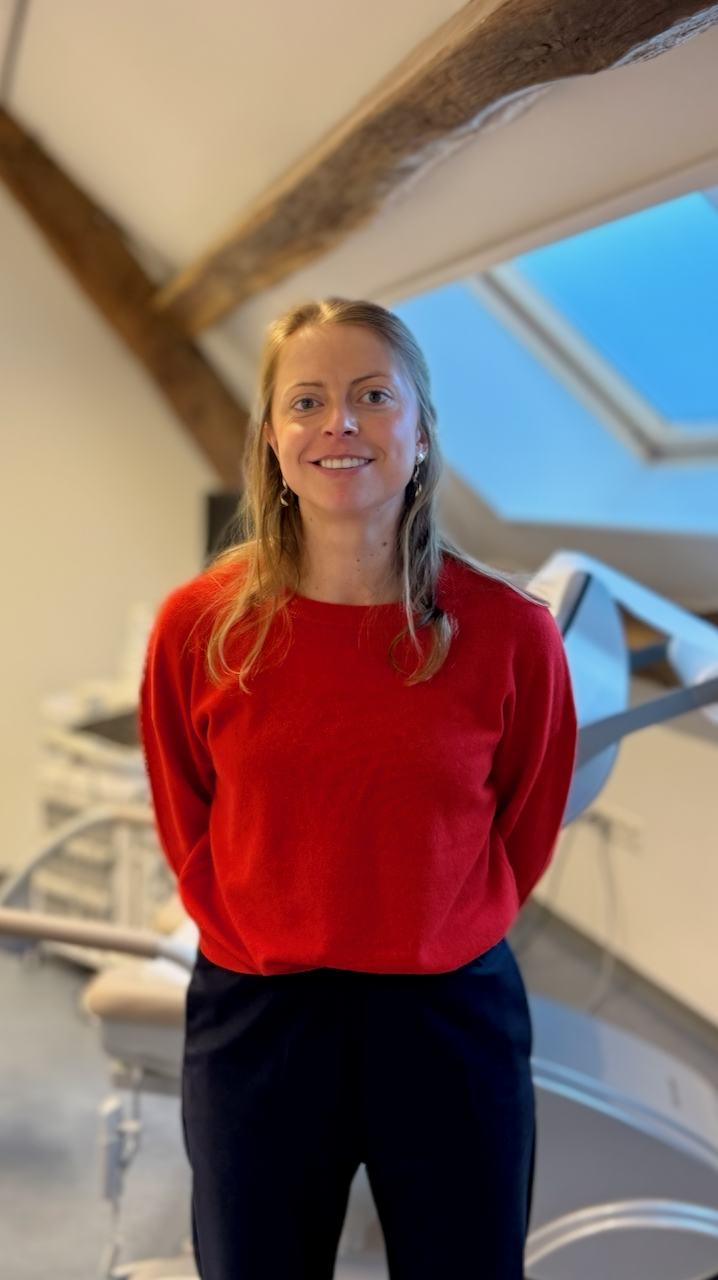 Vrouw met bruin haar in een rode trui en zwarte broek, lachend, in een kamer met een artsenstoel en een dakraam.