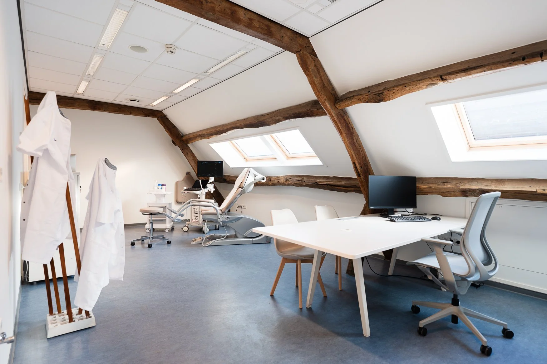 Een medische praktijkruimte met dokterskleding, een doktersstoel, controleapparatuur, een bureau met computer en een stoel, onder een schuin plafond met houten balken en dakramen.