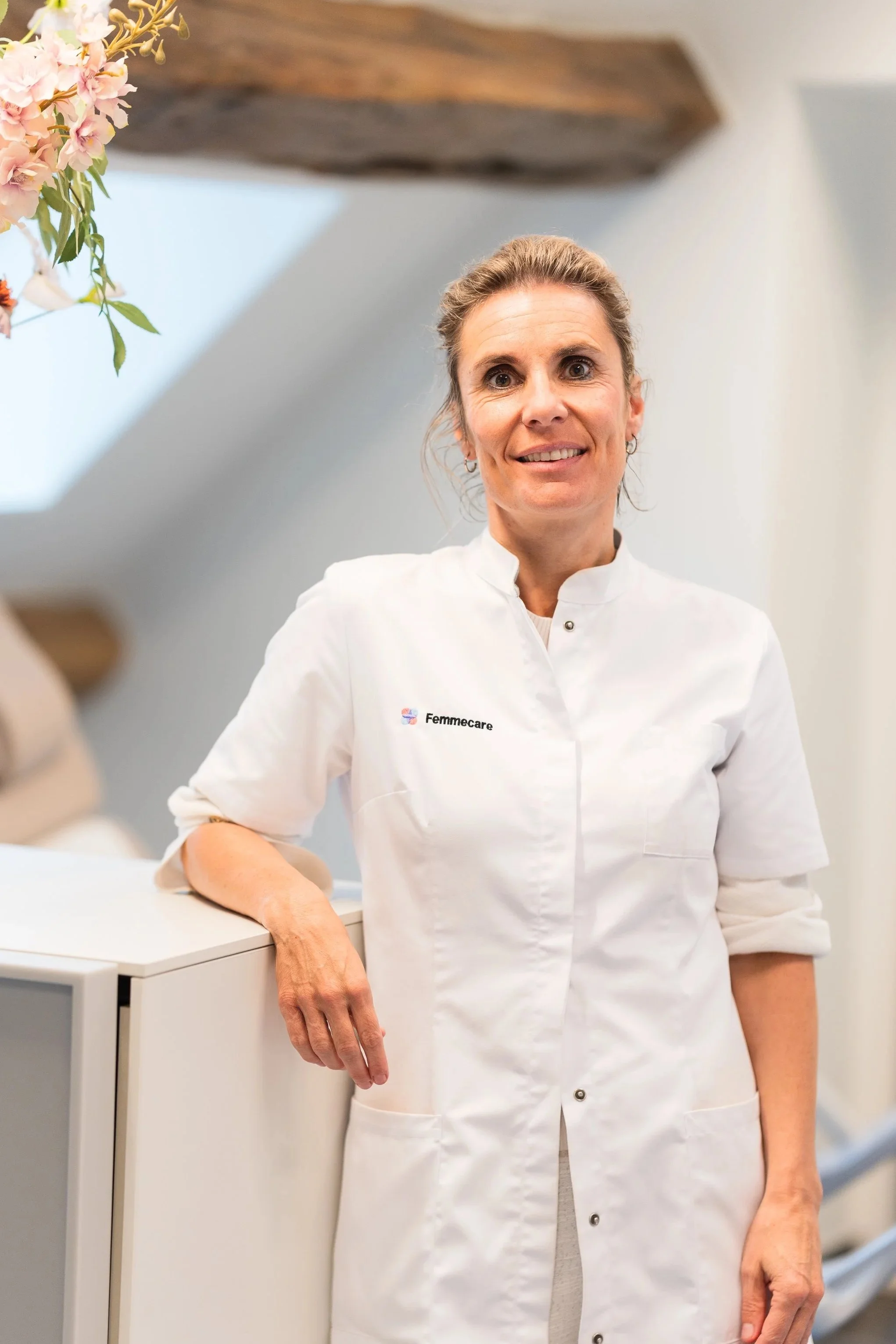 Een vrouw in een witte medische jas met het logo 'Femmecare' staat naast een kastje in een klinische omgeving, met bloemen aan de linkerzijde.