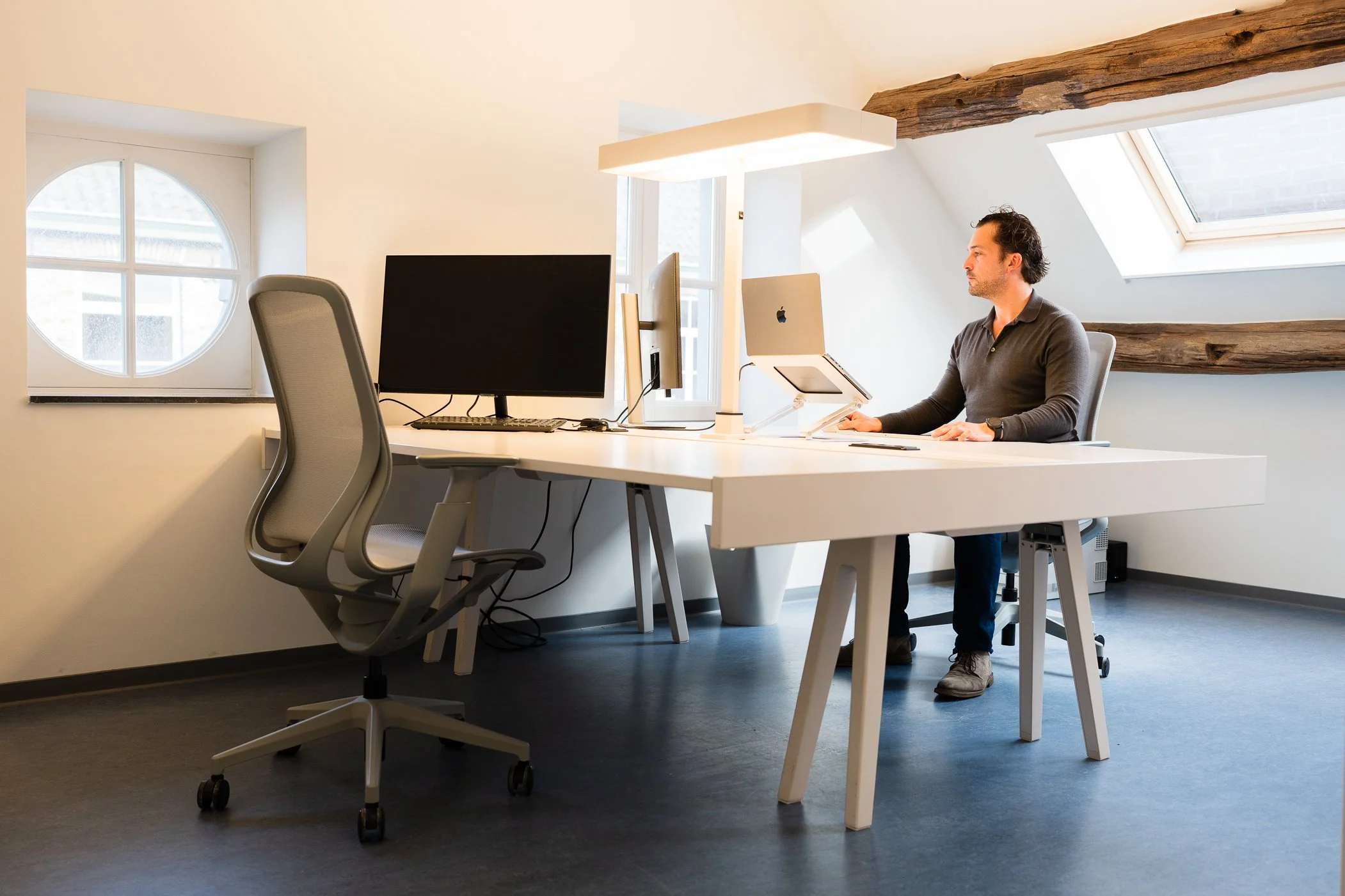 Man zit aan een wit bureau met twee beeldschermen, in een lichte kamer met houten balken en een dakraam.