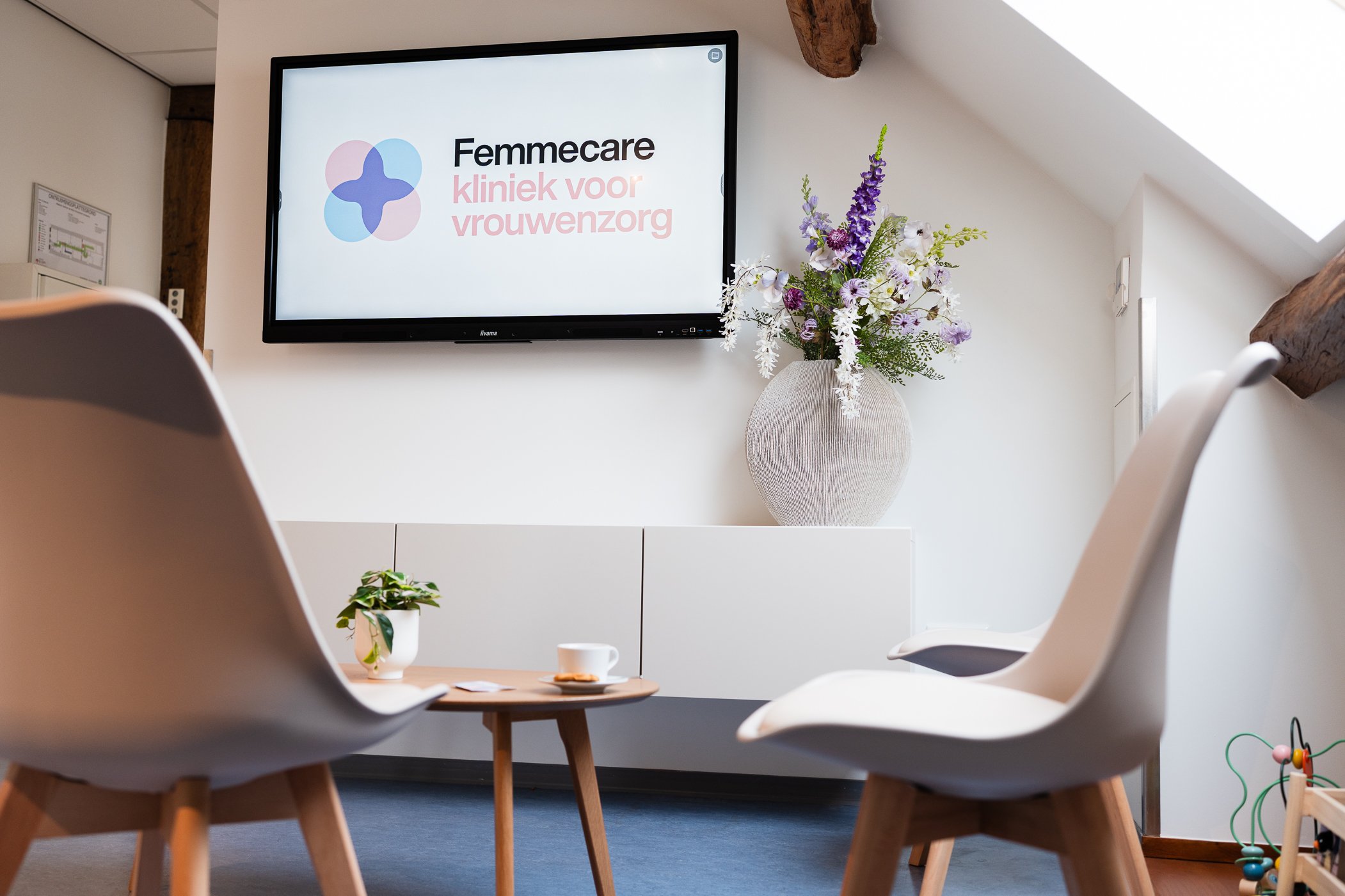 Een overzicht van een wachtkamer of spreekkamer met een muurtelevisie die het logo en slogan van Femacare toont, een grote vaas met bloemen, twee witte stoelen en een kleine houten tafel met een plant en een kopje thee of koffie.