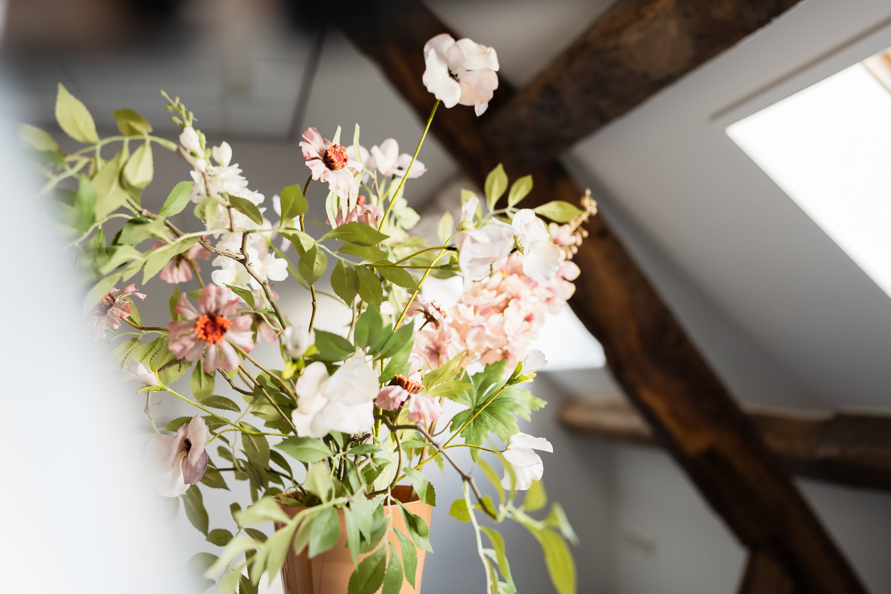 Een bloemenvaas met roze en witte bloemen op een houten surface onder een houten balk, bij een raam.
