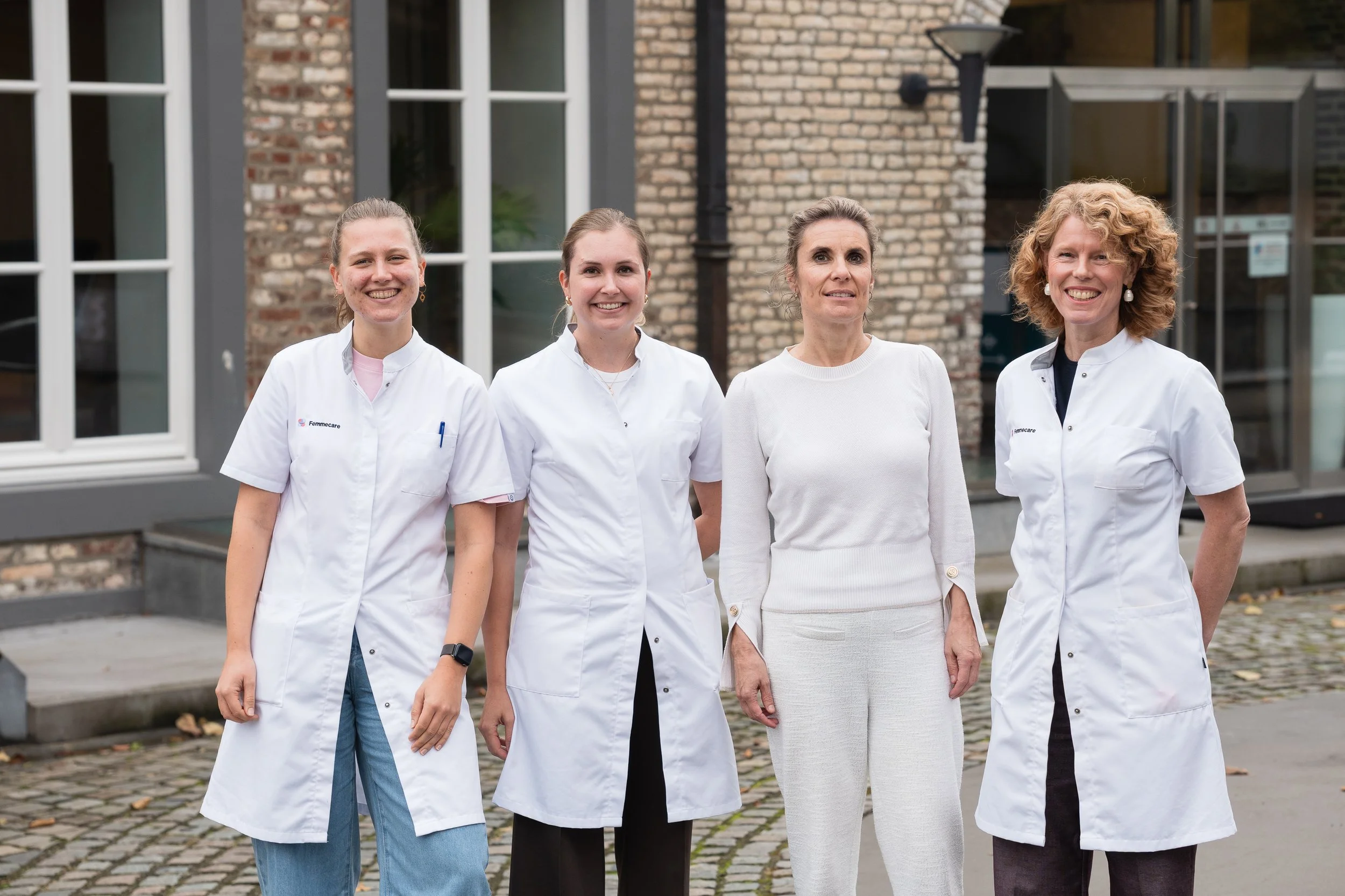 Vijf vrouwen staan buiten voor een gebouw, vier vrouwen dragen witte medische kleding en één vrouw draagt een witte trui, samen poseren ze voor de foto.