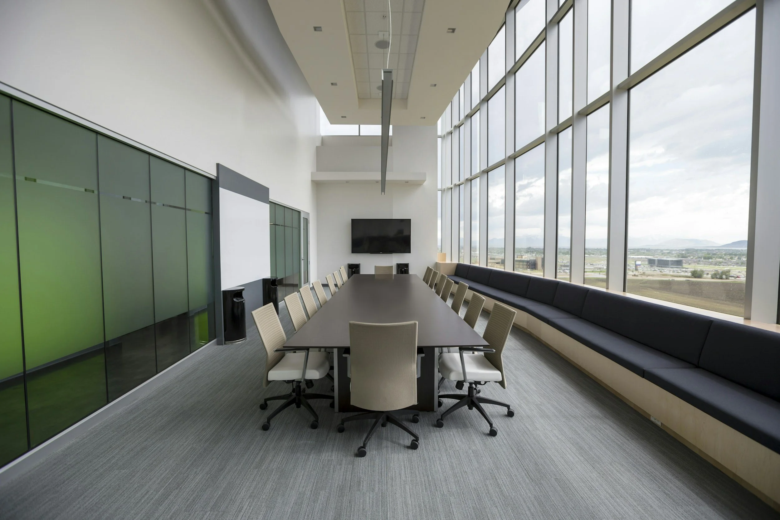 Modern conference room with a long table, beige chairs, large windows with city view, black bench seating along the window, TV on the wall, and a whiteboard.