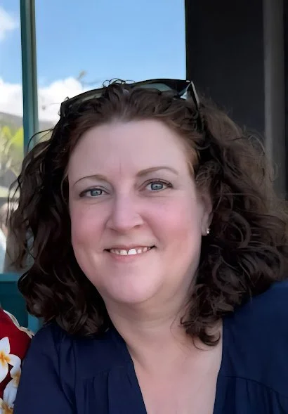 A woman with curly brown hair, light skin, wearing sunglasses on her head, smiling in front of a window with a blue sky outside.
