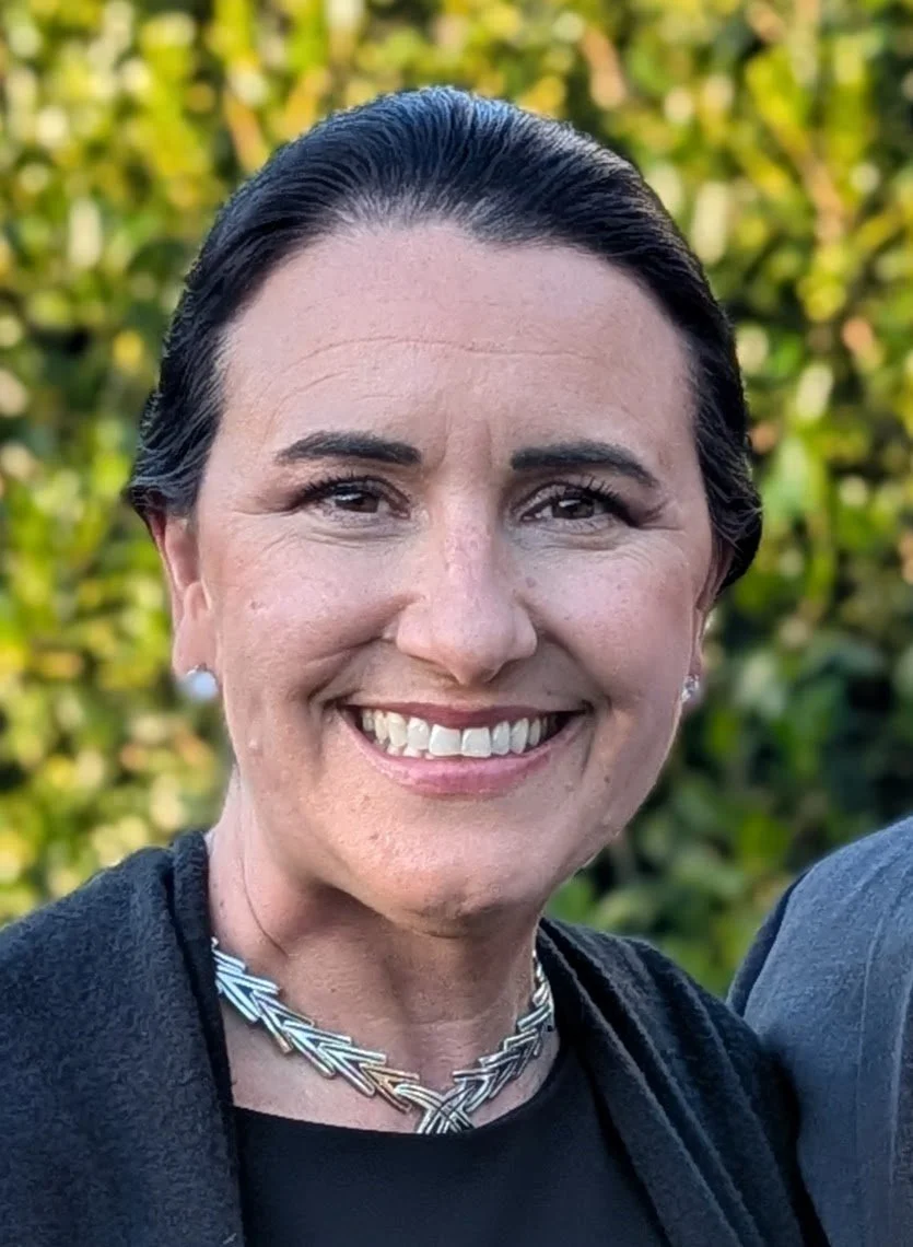 A woman smiling outdoors with greenery in the background.