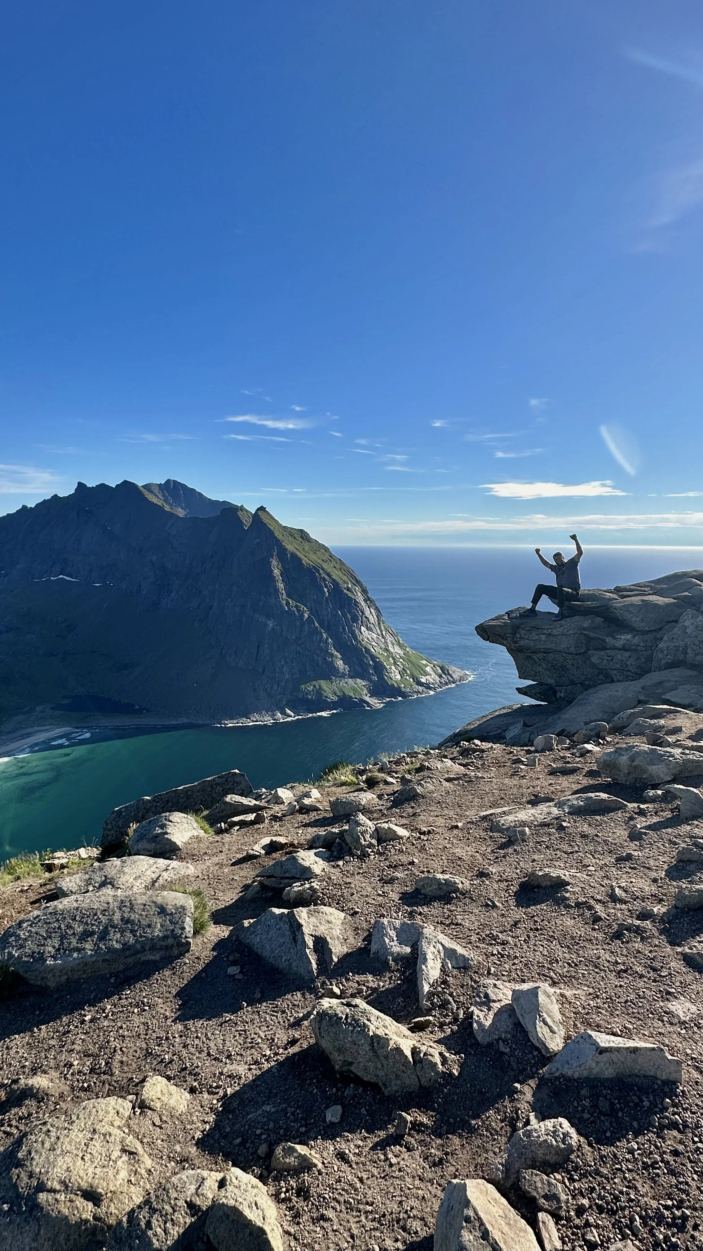 A person sitting on a rock ledge with arms raised in triumph overlooking a fjord and mountains on a clear day.