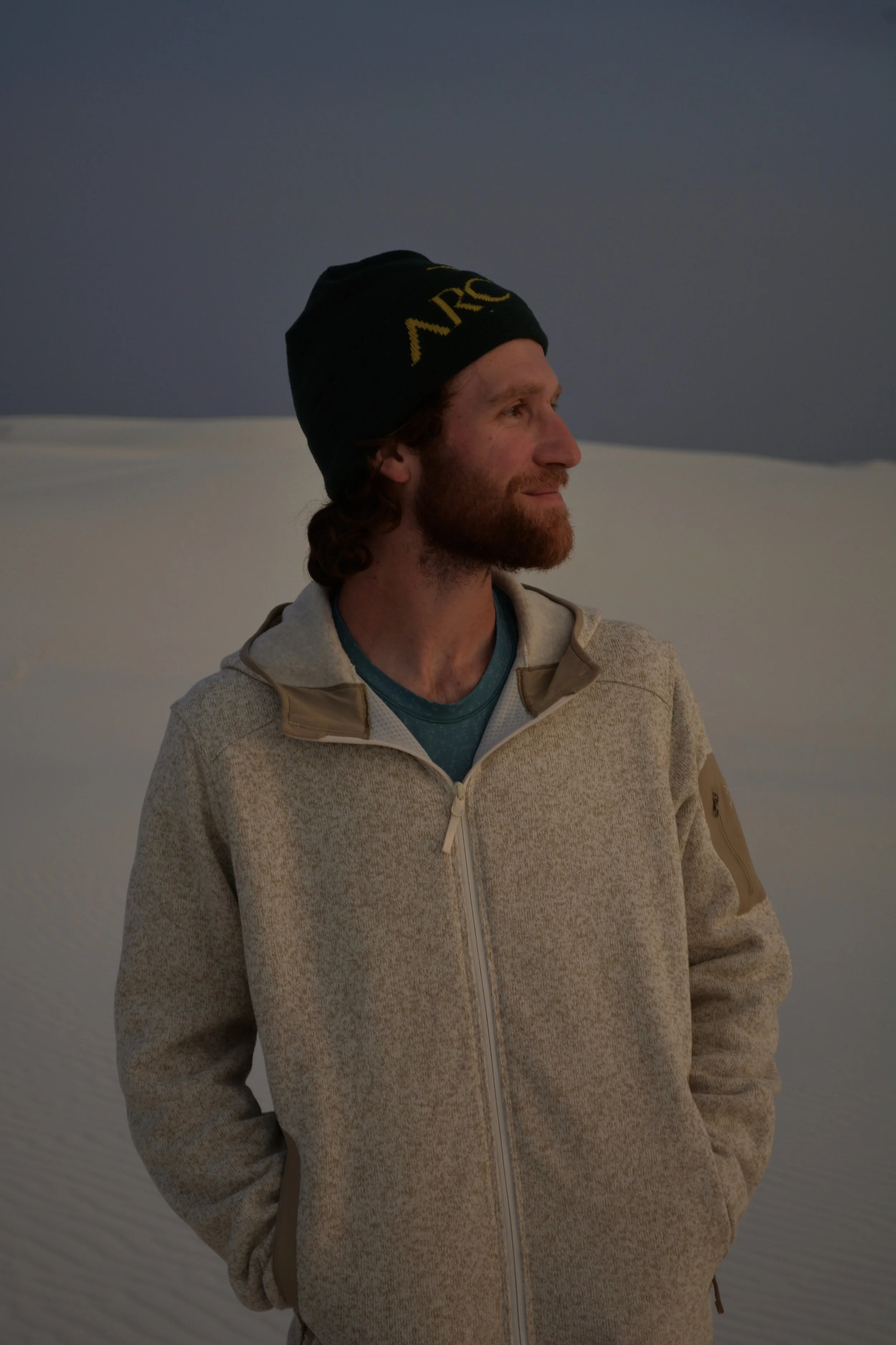 A man with a beard and curly hair wearing a black beanie hat and a beige zip-up jacket stands outdoors against a background of sand dunes and a cloudy sky.