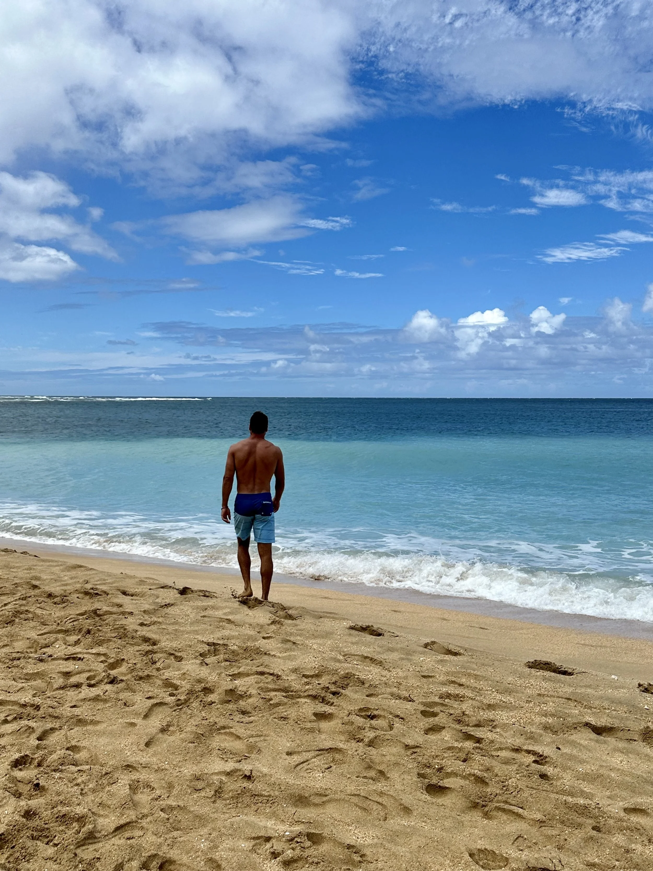 A man in blue swim trunks walking along the sandy beach towards the ocean on a partly cloudy day.